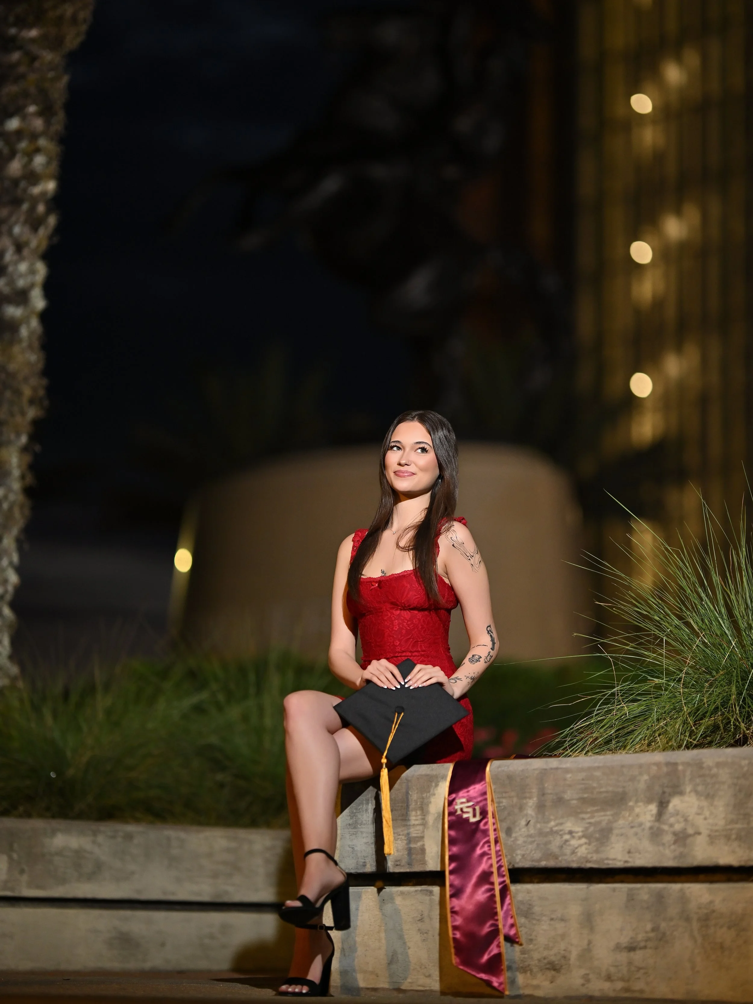 A young woman in a red dress is sitting on a step holding her graduation cap, with a stole hanging from the step. She has tattoos on her arms and is wearing black heels. The background features a large building with lit windows and some bushes.