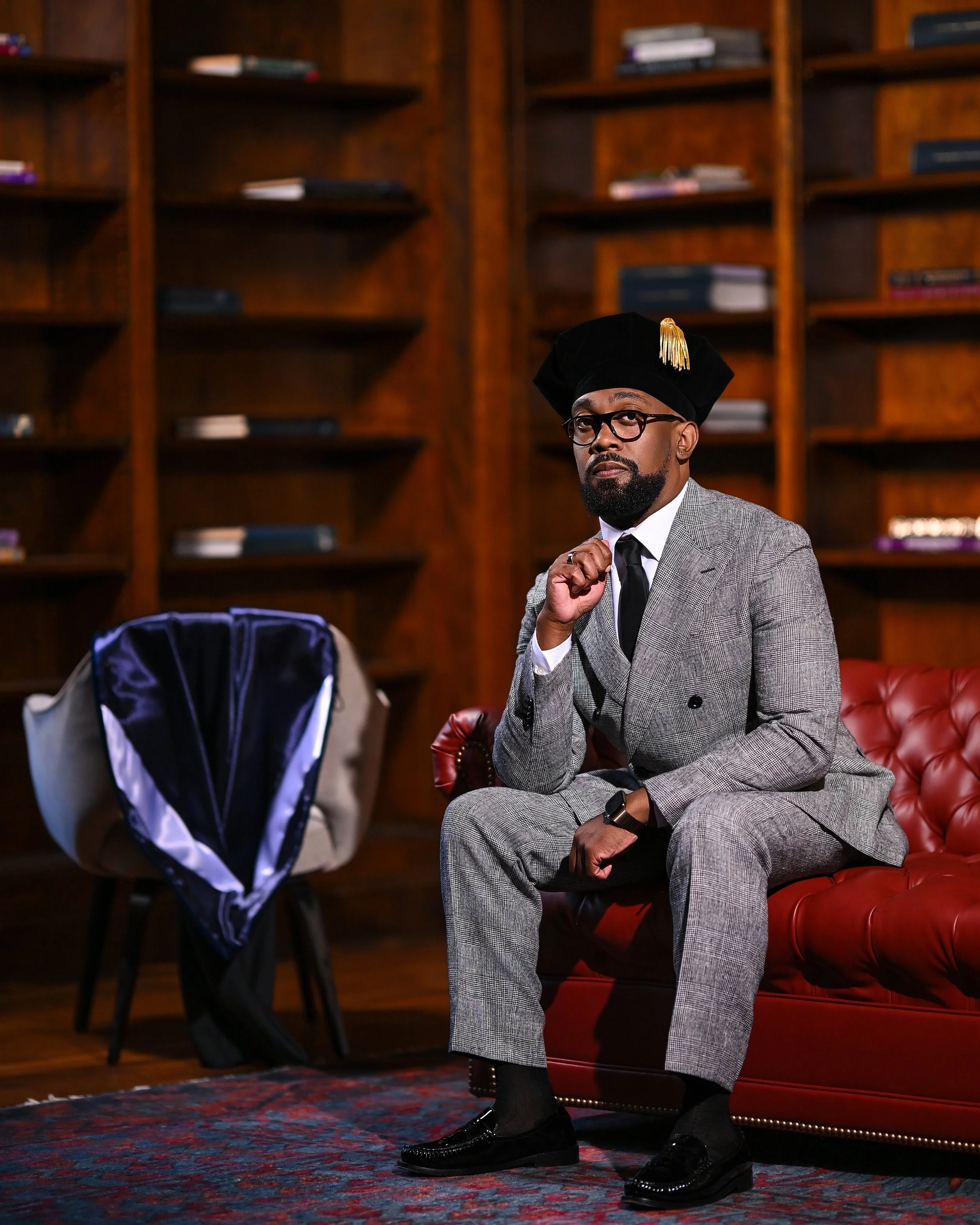 A man in a gray plaid suit, black tie, and glasses sitting on a red leather sofa in a room with wooden bookshelves. He is wearing a black academic cap with a gold tassel and has a contemplative expression. A blue backpack is in the foreground, and bo