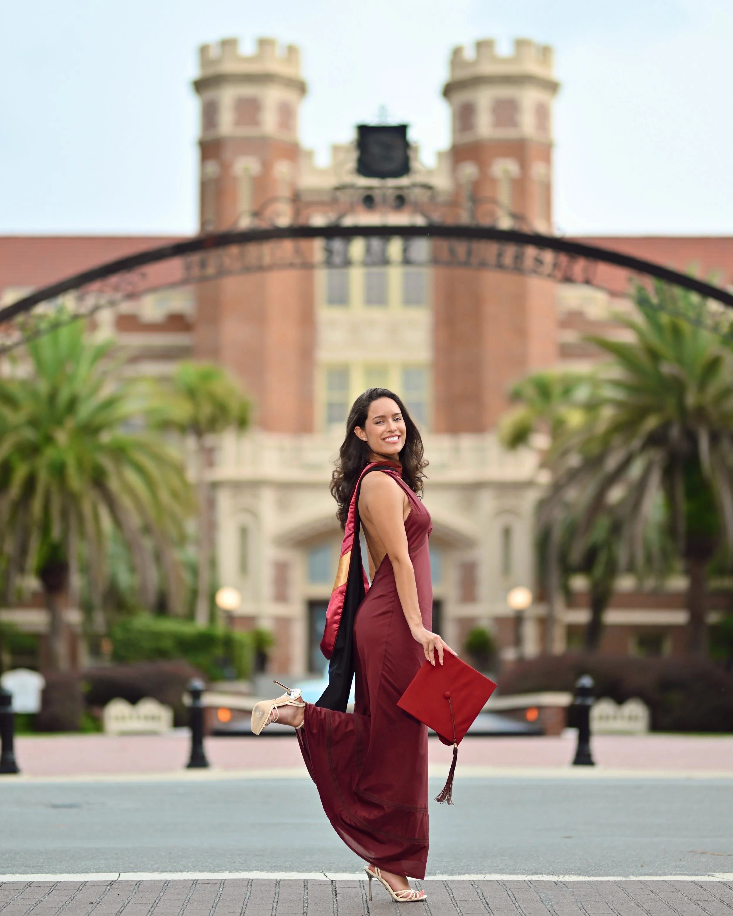A young woman in a burgundy dress is jumping in the air while holding a graduation cap in her right hand. She is smiling and has a shoulder bag. The background shows a historic brick building with two towers and a clock, with green trees on either si