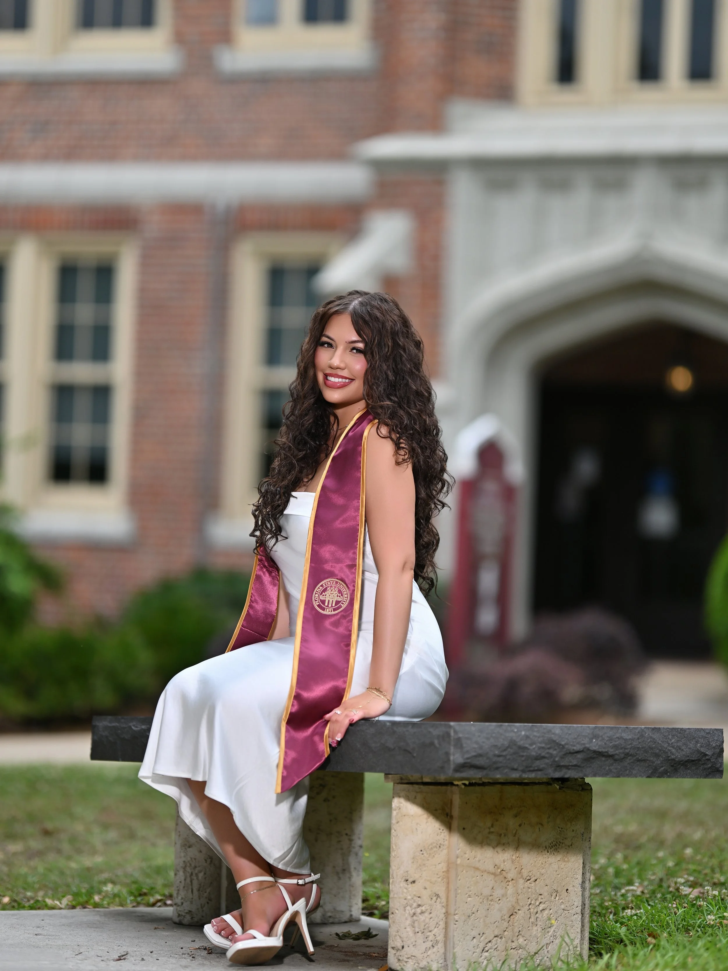 A young woman in a white dress and high heels is sitting on a stone bench in front of a red-brick building. She is wearing a graduation stole and looks happy.