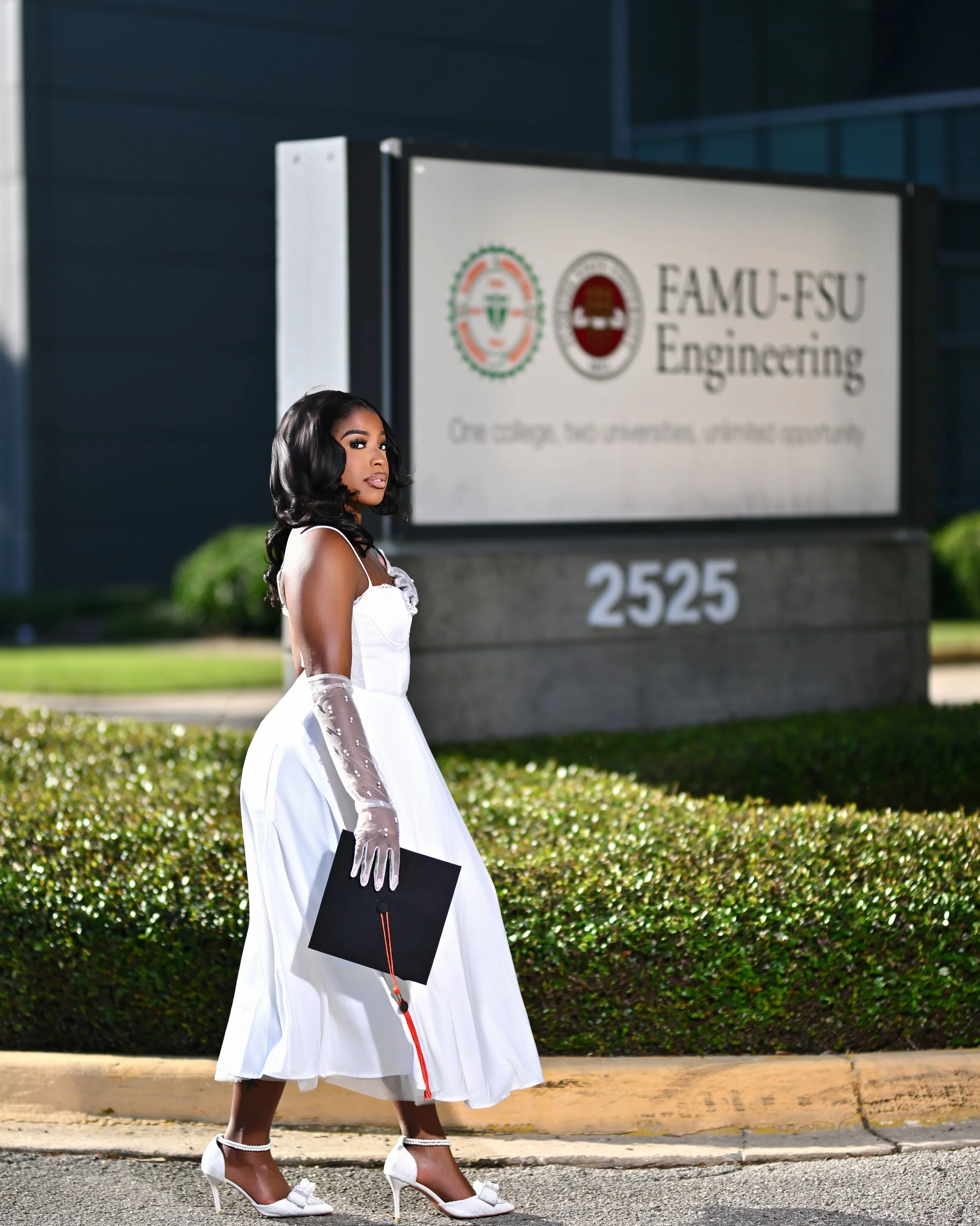 Young woman in a white dress with sheer gloves and high heels holding a diploma outside on a sidewalk next to a hedge, with a sign for FAMU-FSU Engineering in the background.