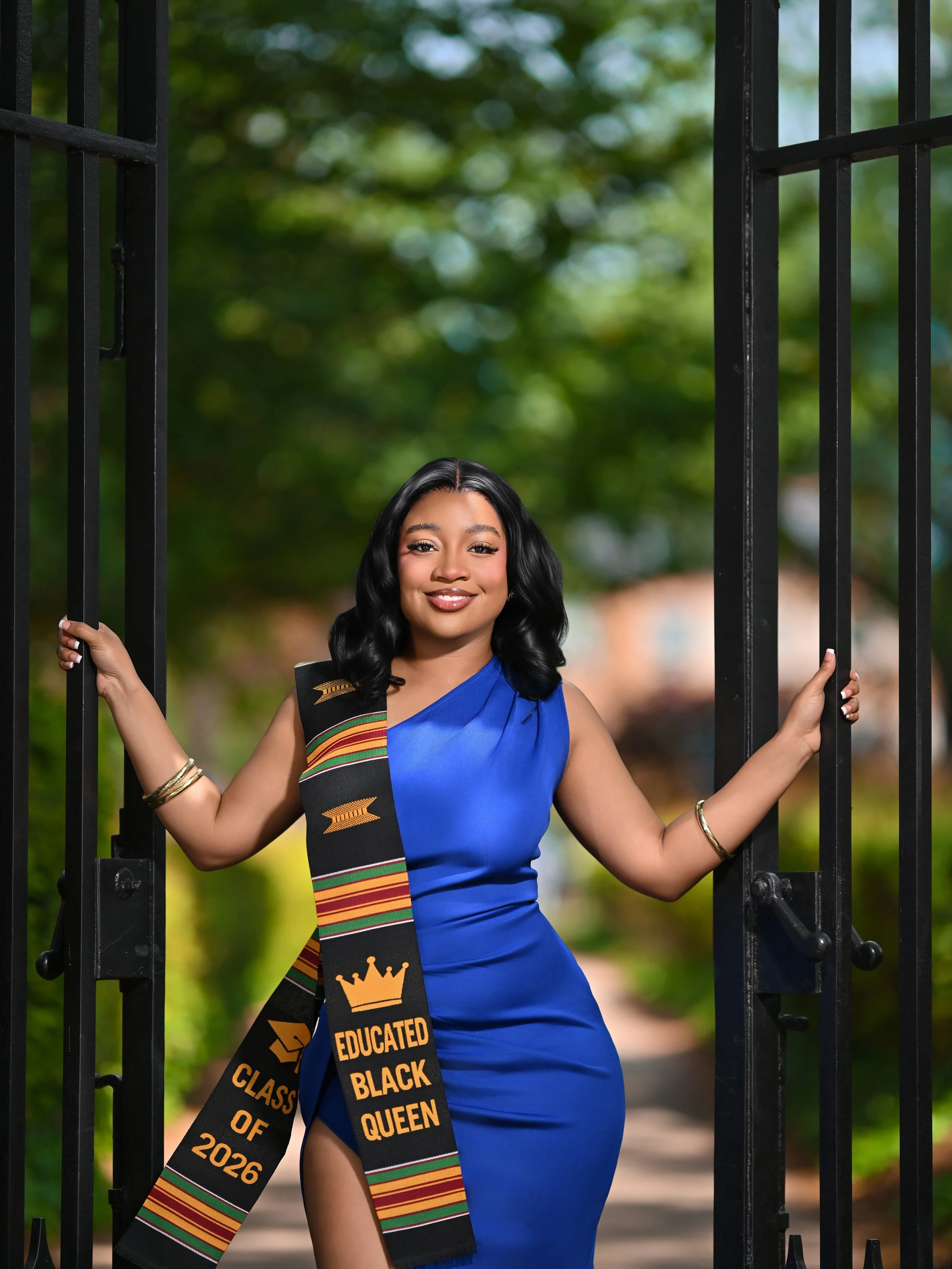 Young Black woman in a blue dress, smiling, wearing a graduation sash that reads 'Class of 2026' and 'Educated Black Queen,' standing in an open gate with green trees in the background.