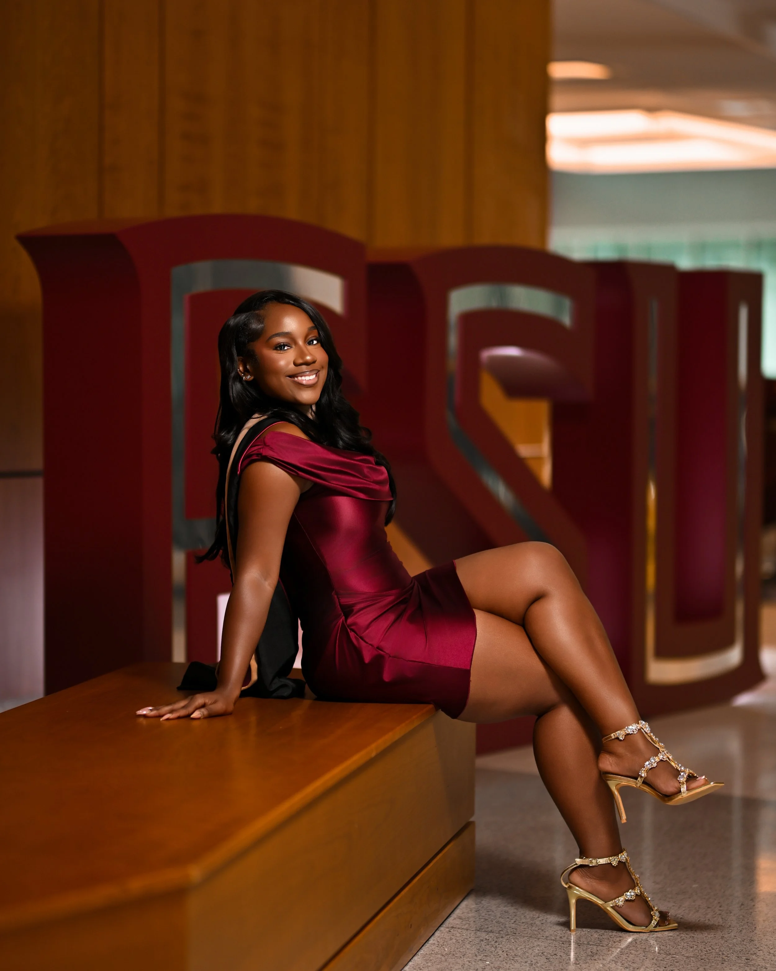 A young woman in a burgundy satin dress with an off-the-shoulder design, sitting on a wooden surface, smiling at the camera, wearing gold high-heeled sandals with rhinestone embellishments.