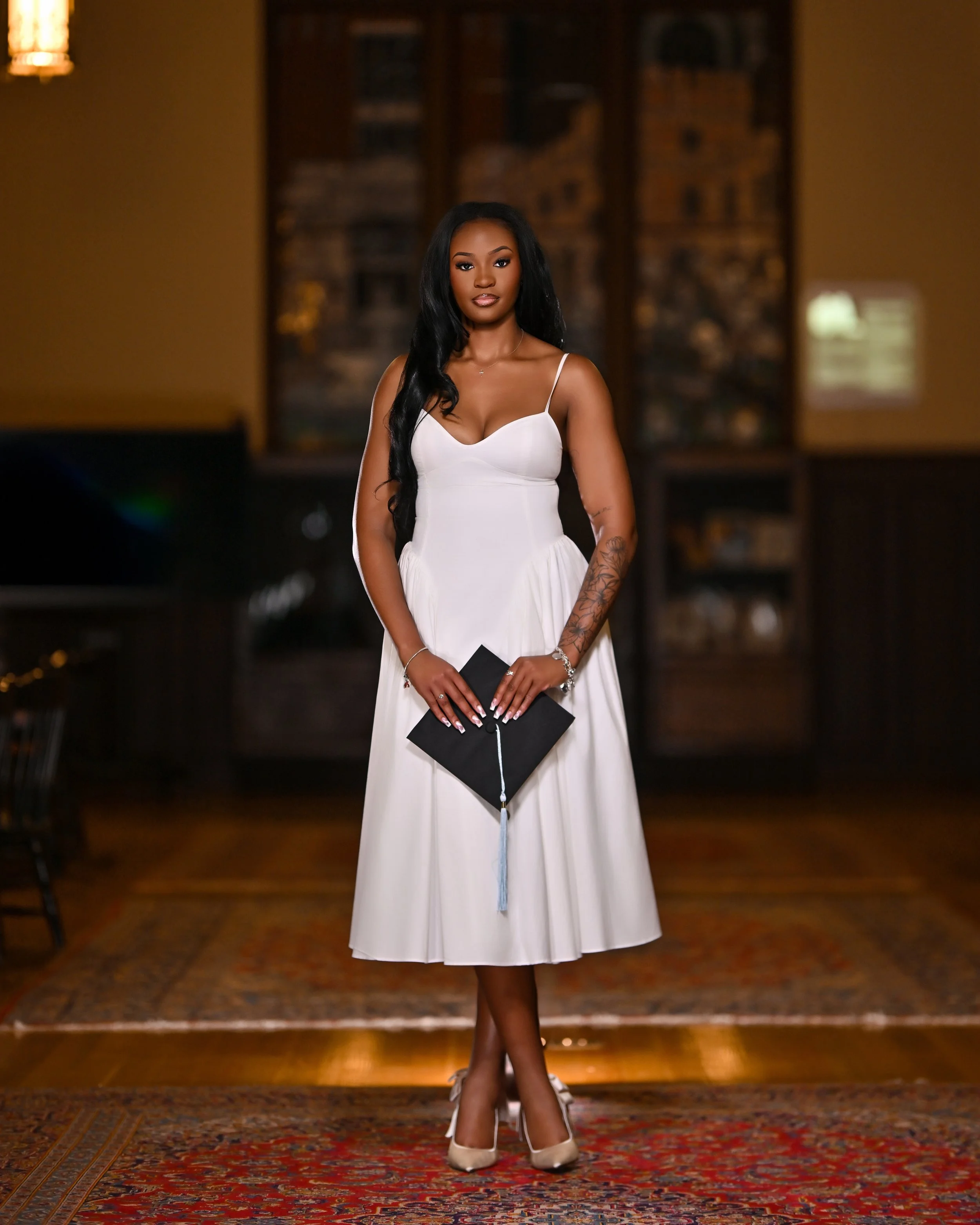 A woman with long black hair wearing a white strapless dress and high heels stands on a patterned carpet, holding a black graduation cap. The background features a blurred brick edifice mural.
