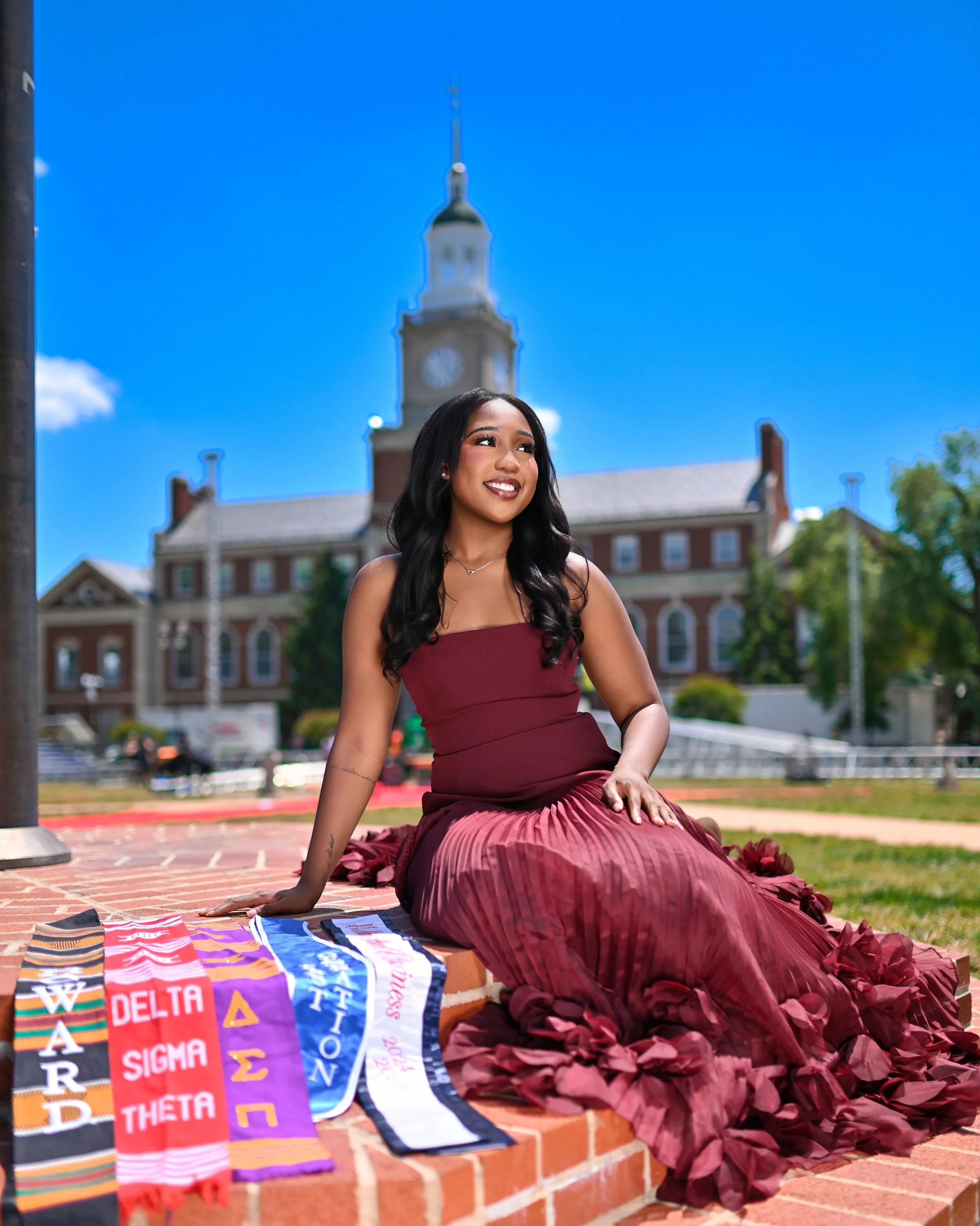 A young woman with long black hair and a maroon dress sitting on a brick ledge outdoors, smiling with a clear blue sky and a clock tower in the background. There are multiple colorful banners and sashes with Greek letters and fraternity names on the 