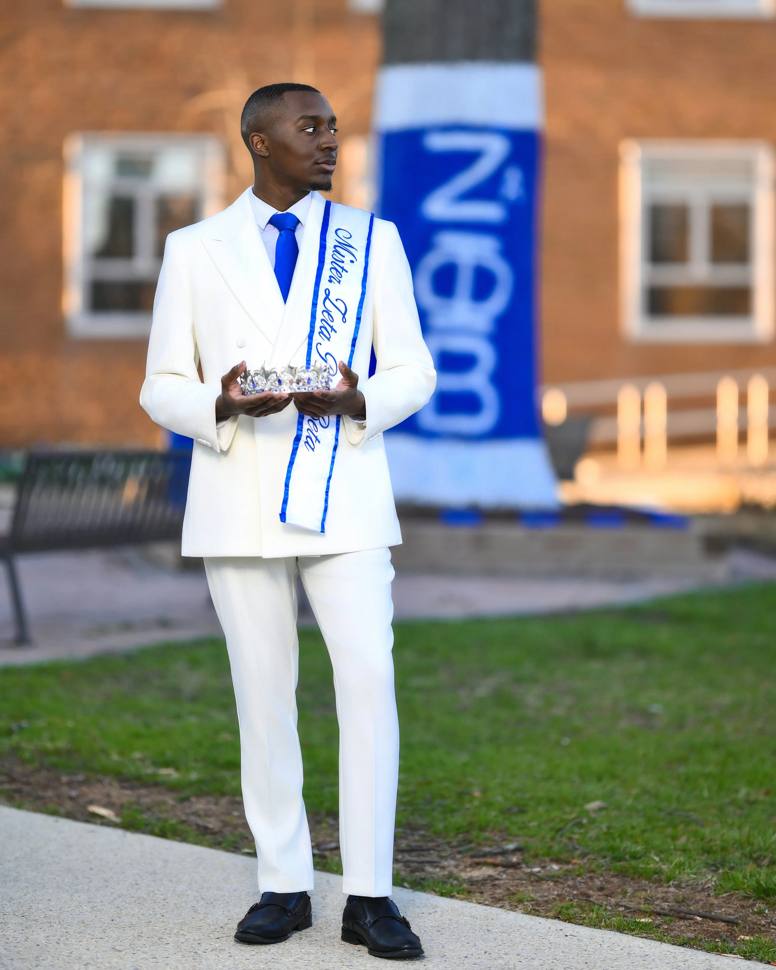 A young man dressed in a white tuxedo with a blue tie and a sash that reads 'Alpha Phi Alpha' is holding a crystal crown in his hands. He is standing outdoors near a building with brick walls and windows, and there is a blue and white banner in the b