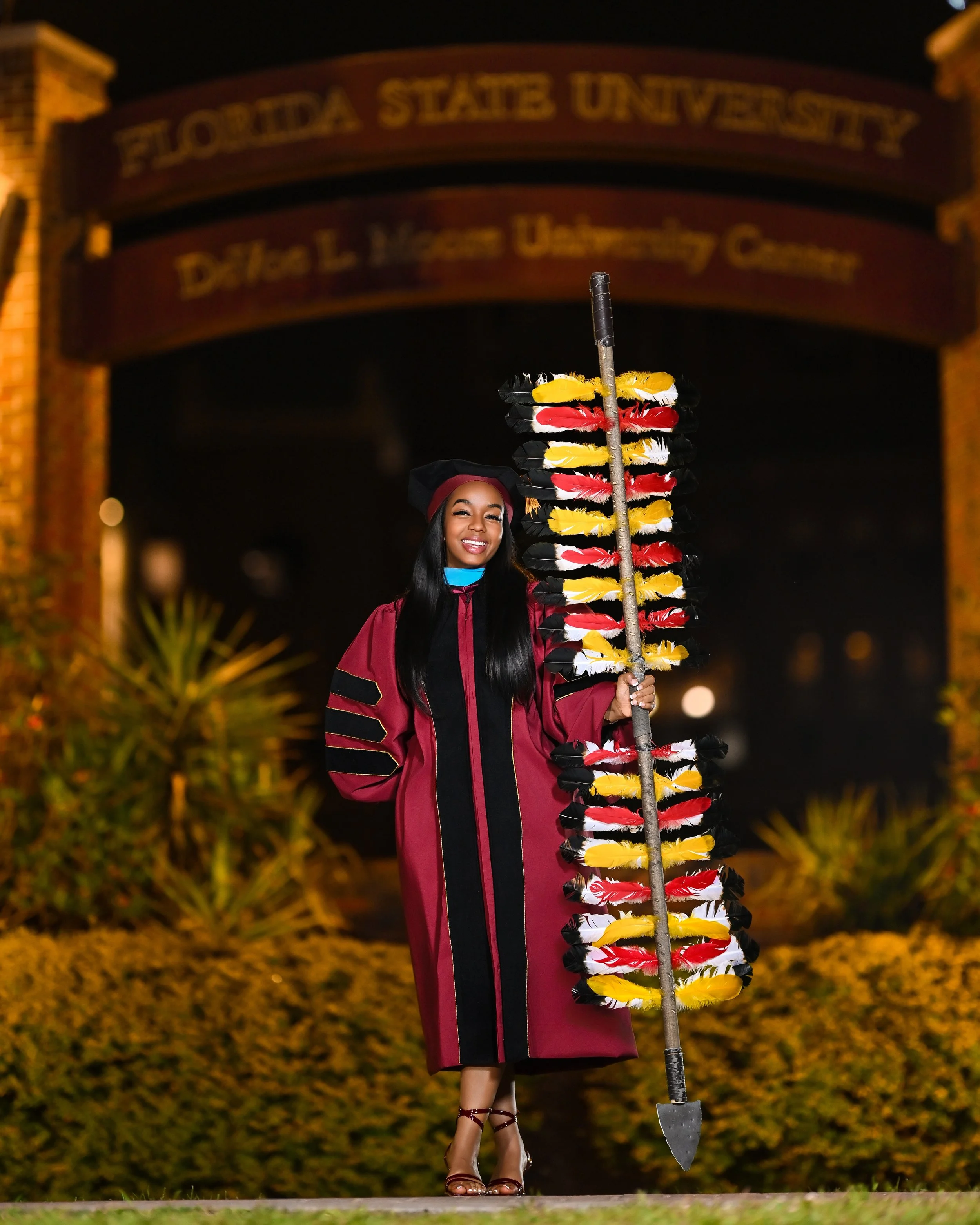 Graduate woman in cap and gown holding a decorated scepter, standing outside at night in front of an arched Florida State University sign.