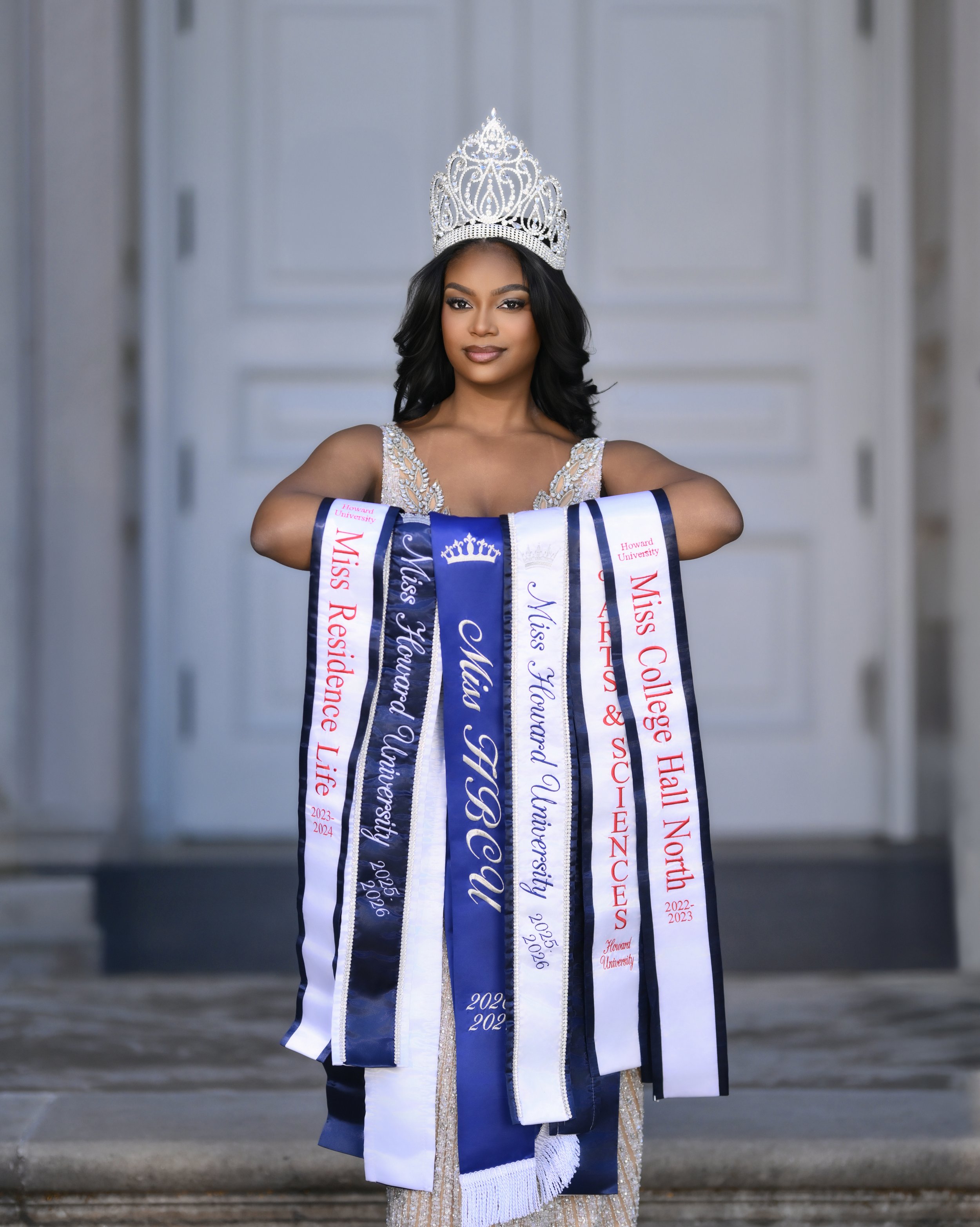A young woman wearing a silver tiara and a sash with multiple ribbons, standing on steps with a gray background. She has dark wavy hair, light makeup, and a confident expression.