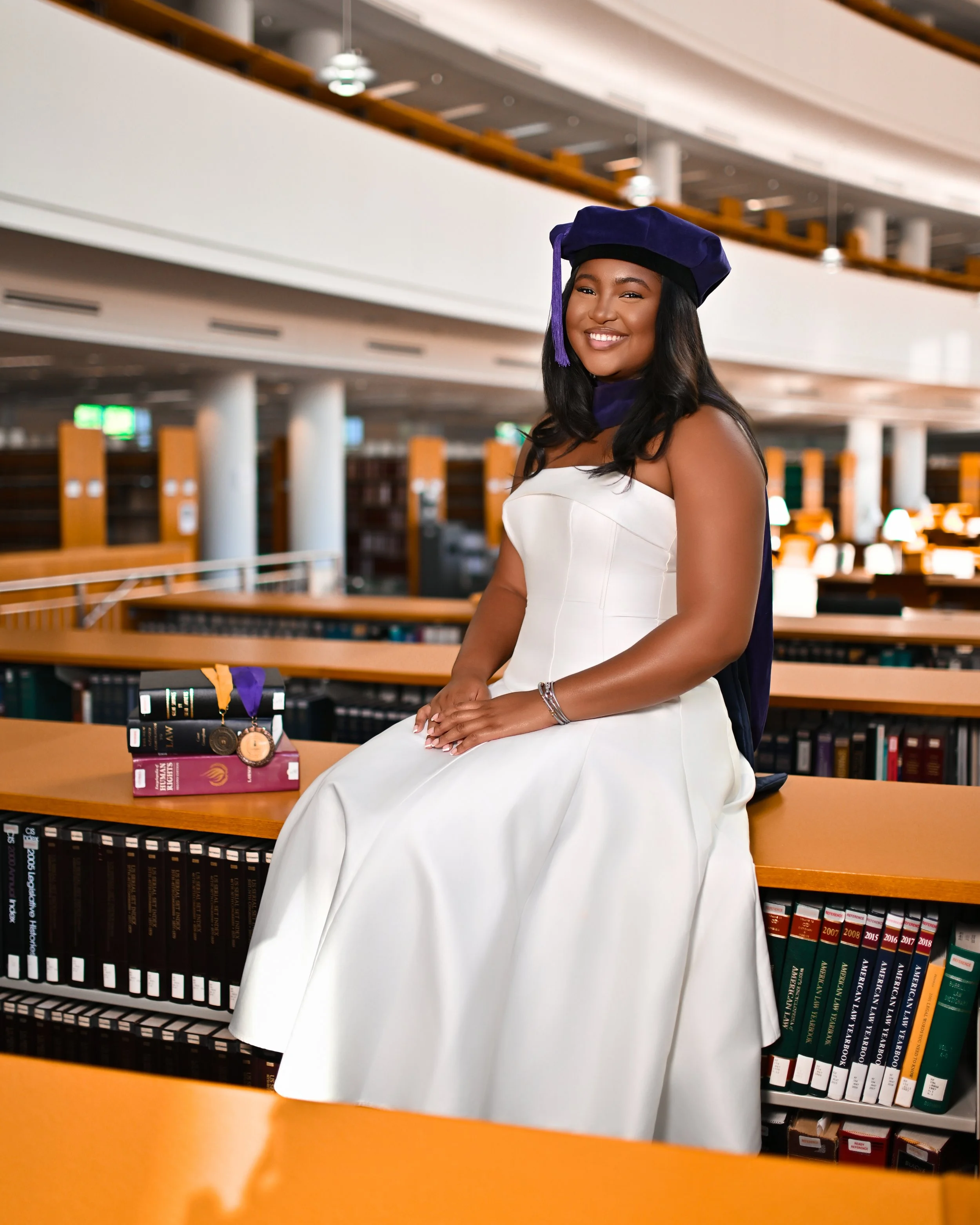 A young woman in a white dress and graduation cap is sitting in a library with bookshelves in the background. There are graduation medals and books on the table beside her.