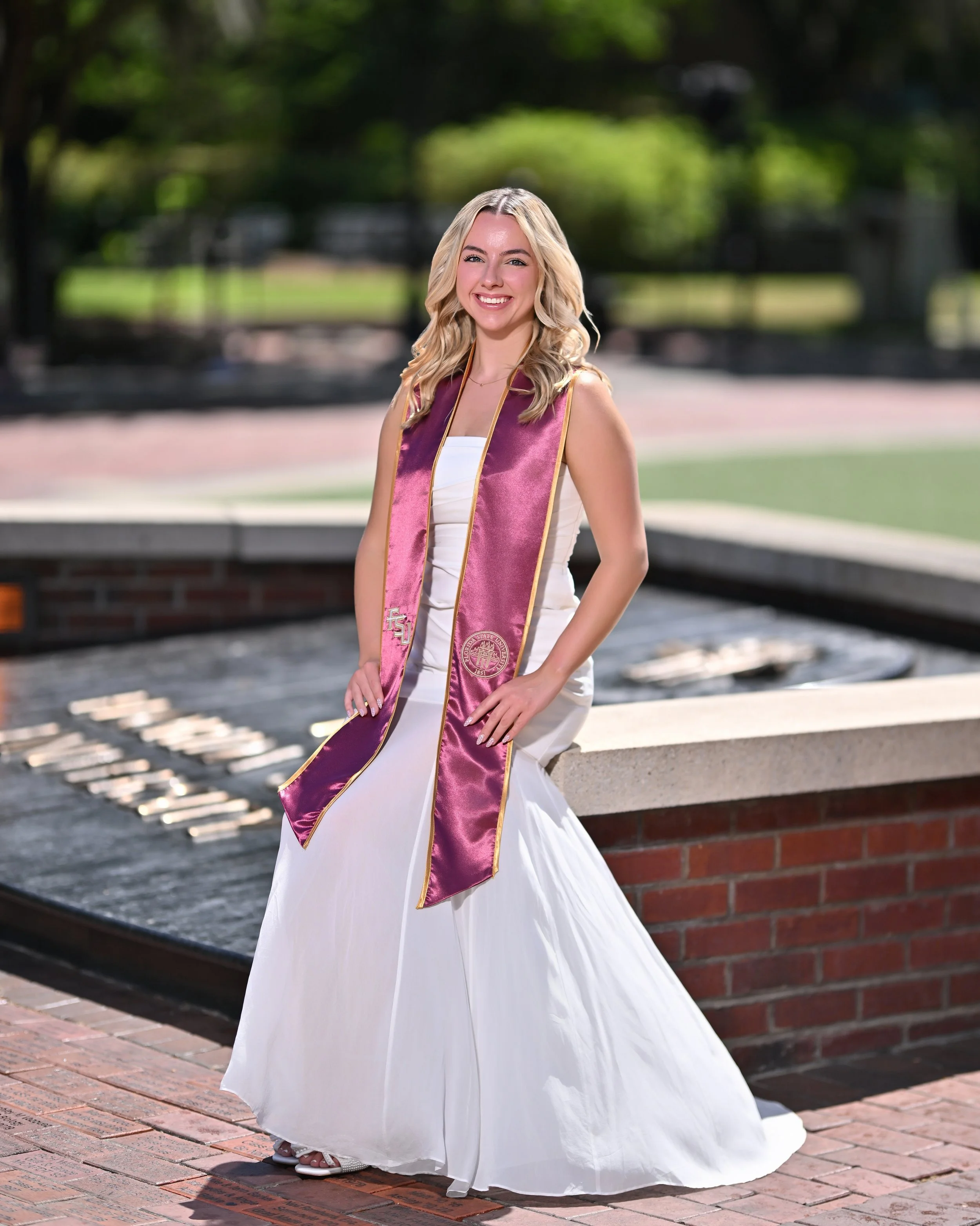 Young woman in a white dress wearing a maroon and gold graduation stole, smiling standing outdoors on a brick pathway with greenery in the background.