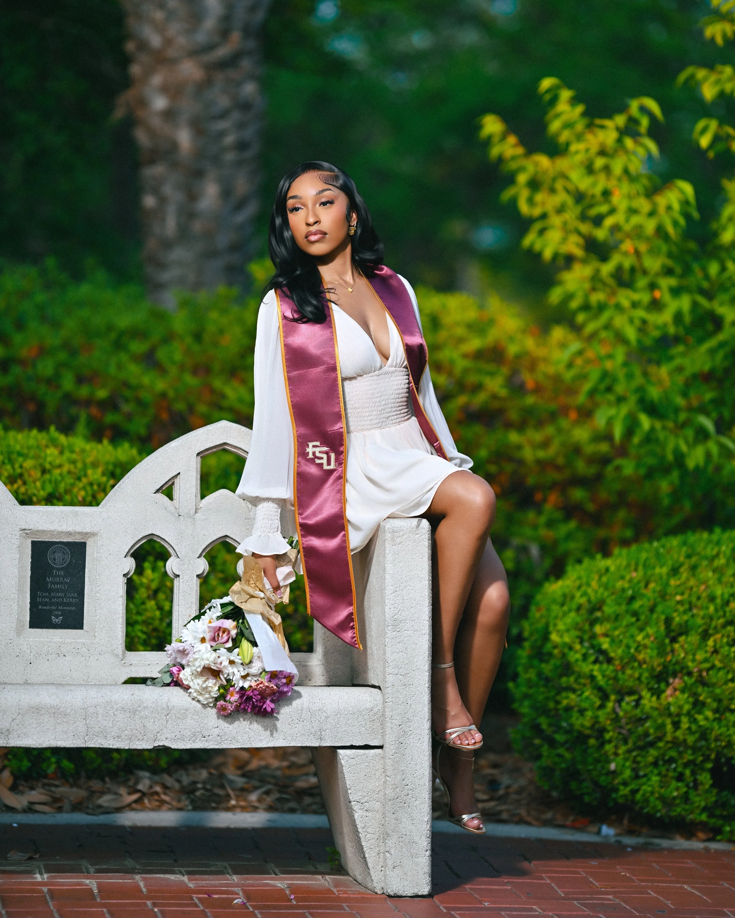 Young woman in a white dress and maroon graduation stole sitting on a white bench, outdoors with green foliage and a palm tree in the background, holding a bouquet of flowers.