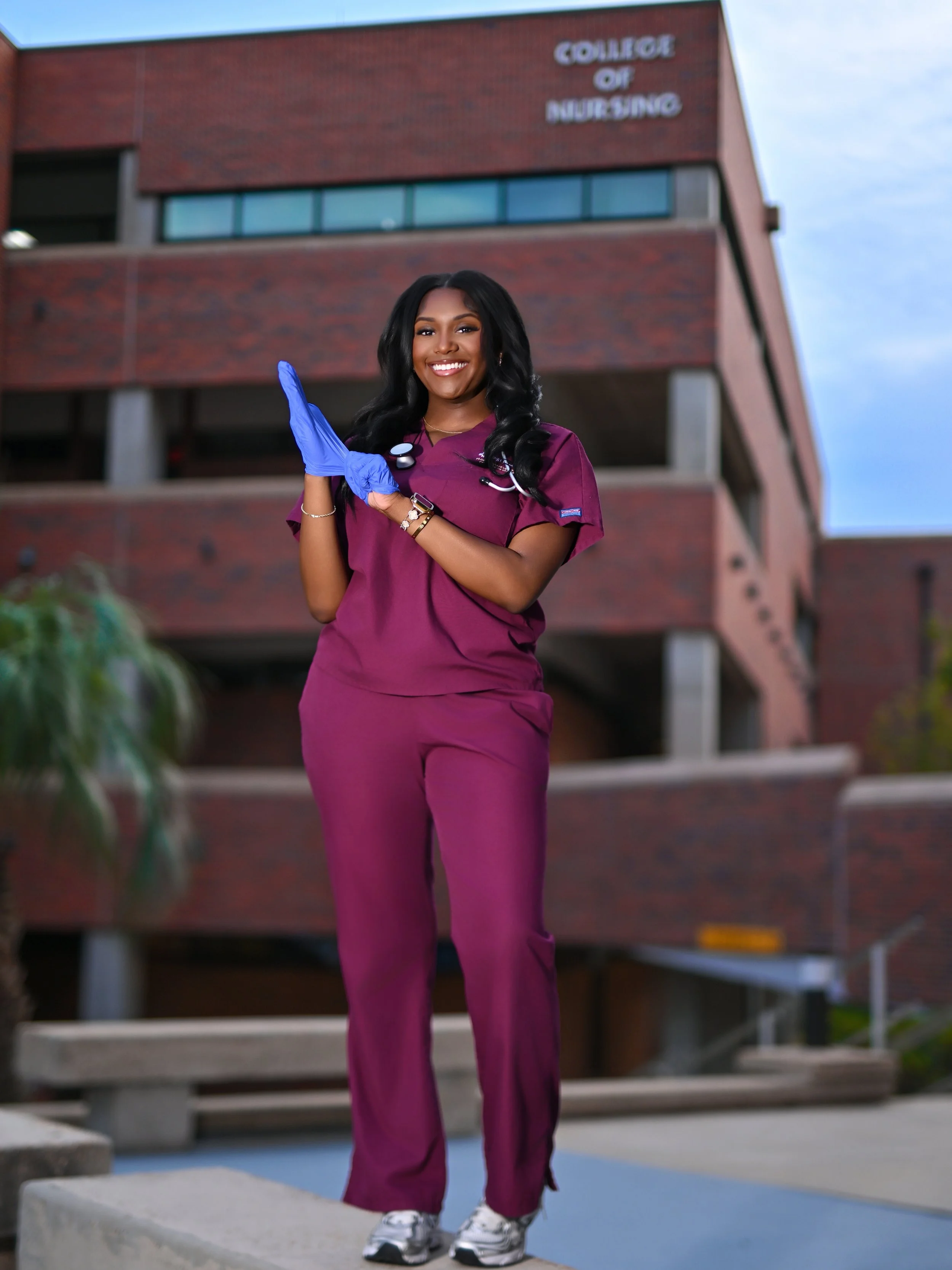 A smiling woman wearing medical scrubs and gloves, standing outdoors in front of a building with the sign 'College of Nursing'.