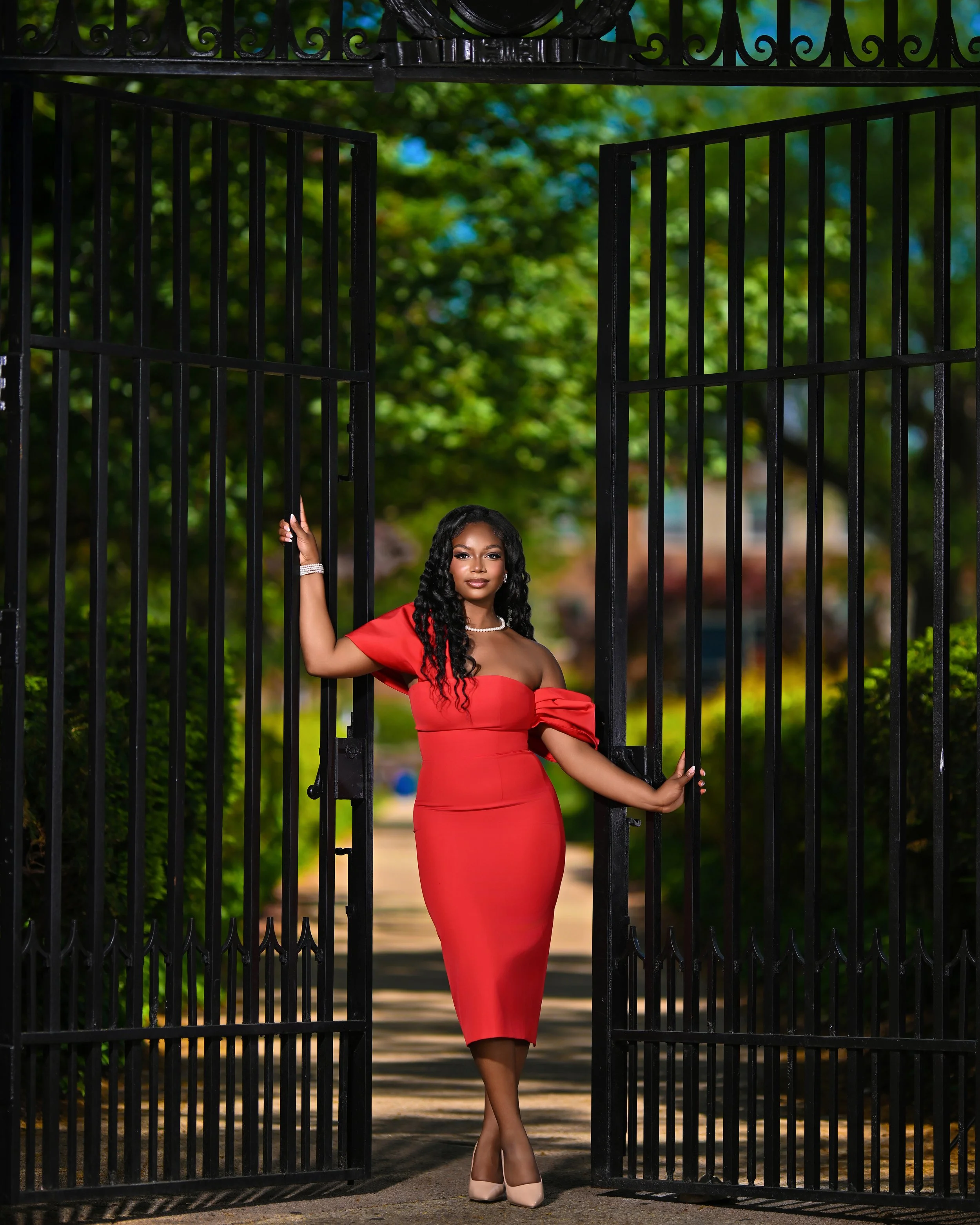 A woman in a red dress and beige heels standing at an open black gate, with green trees in the background.