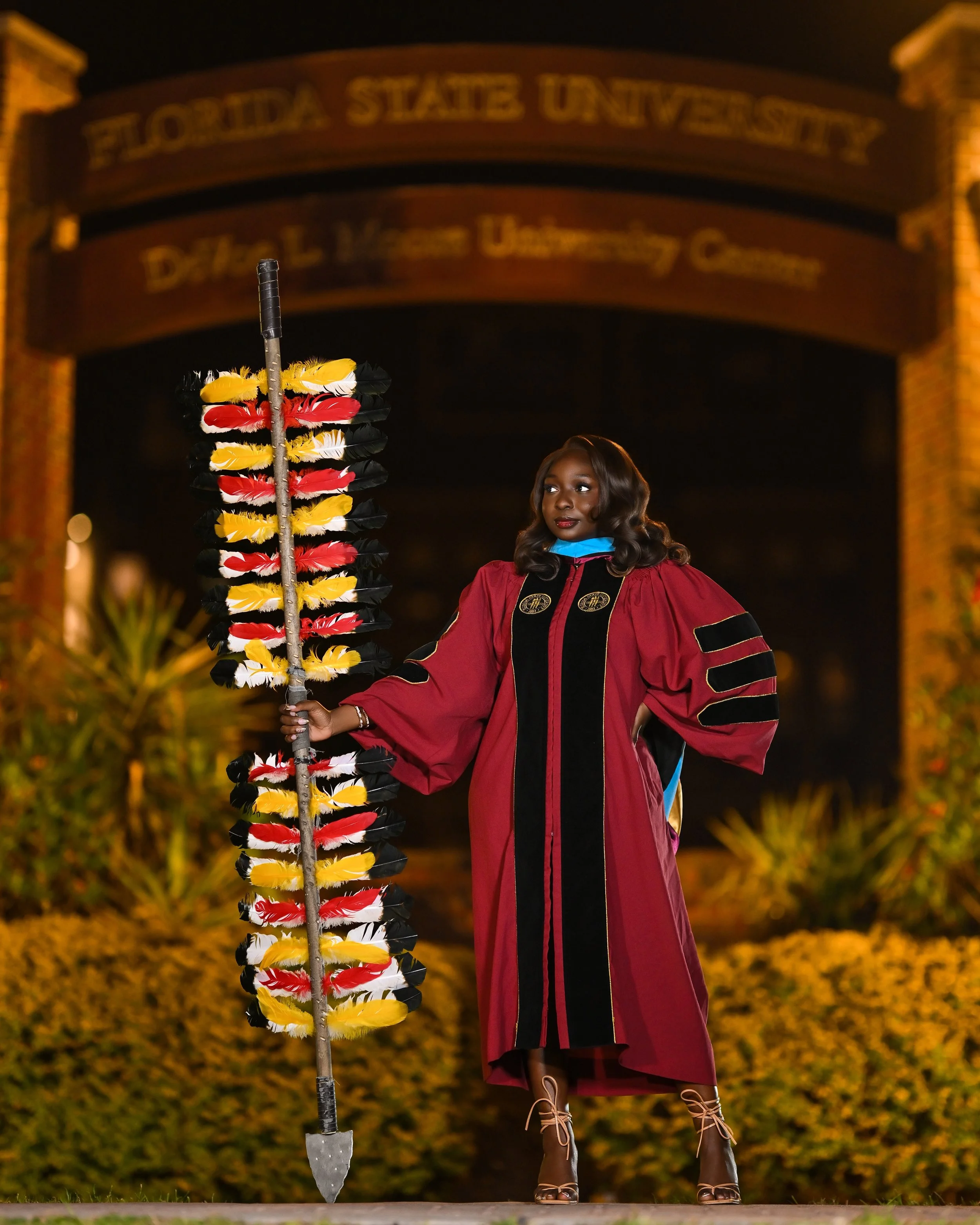 Graduate woman in maroon cap and gown holding a colorful feathered staff at night, with the Florida State University sign in the background.