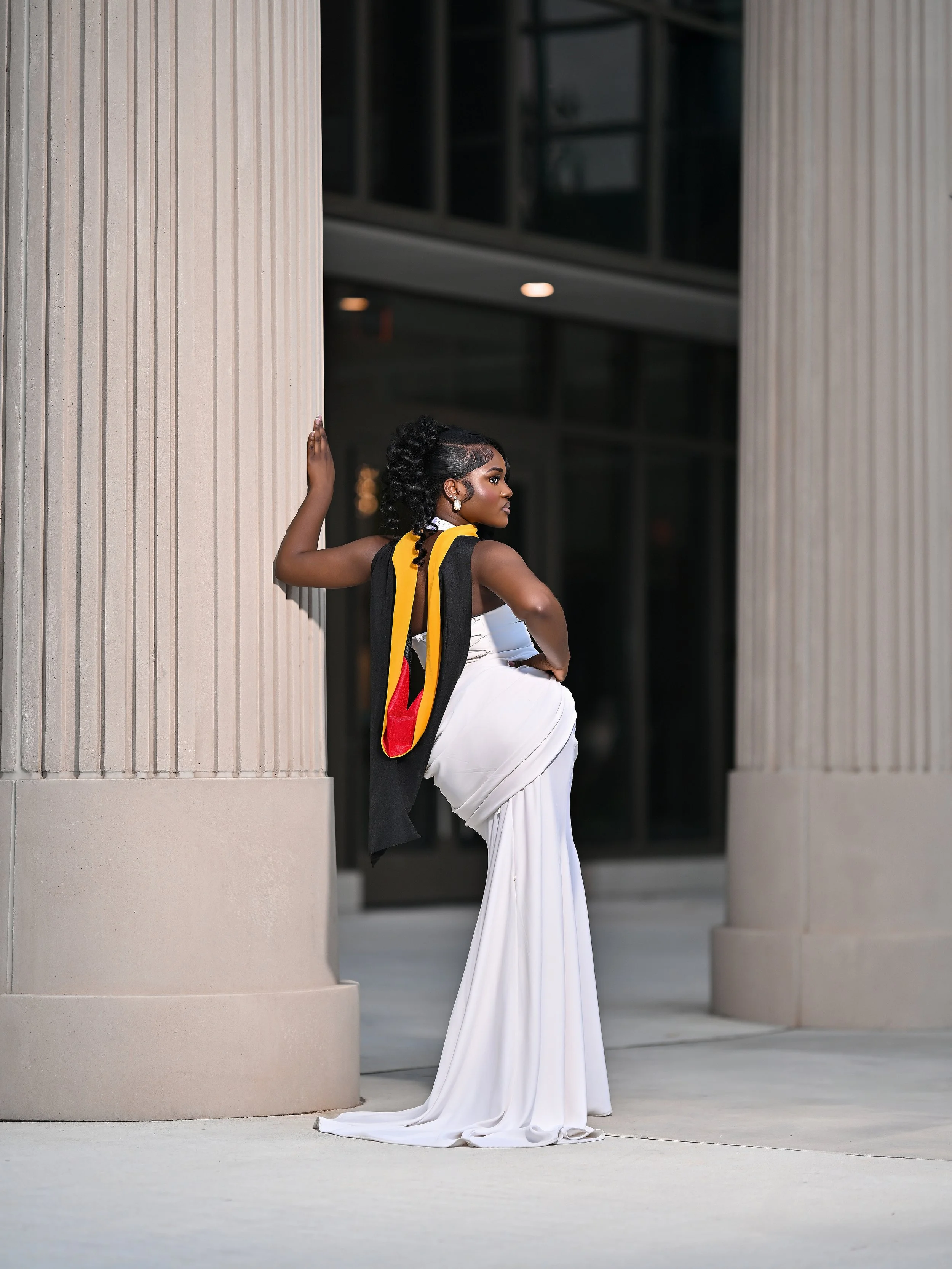 A woman in a white dress with black and yellow accents, standing between large pillars, looking to her left, with her right hand touching the pillar, and her left hand on her waist.
