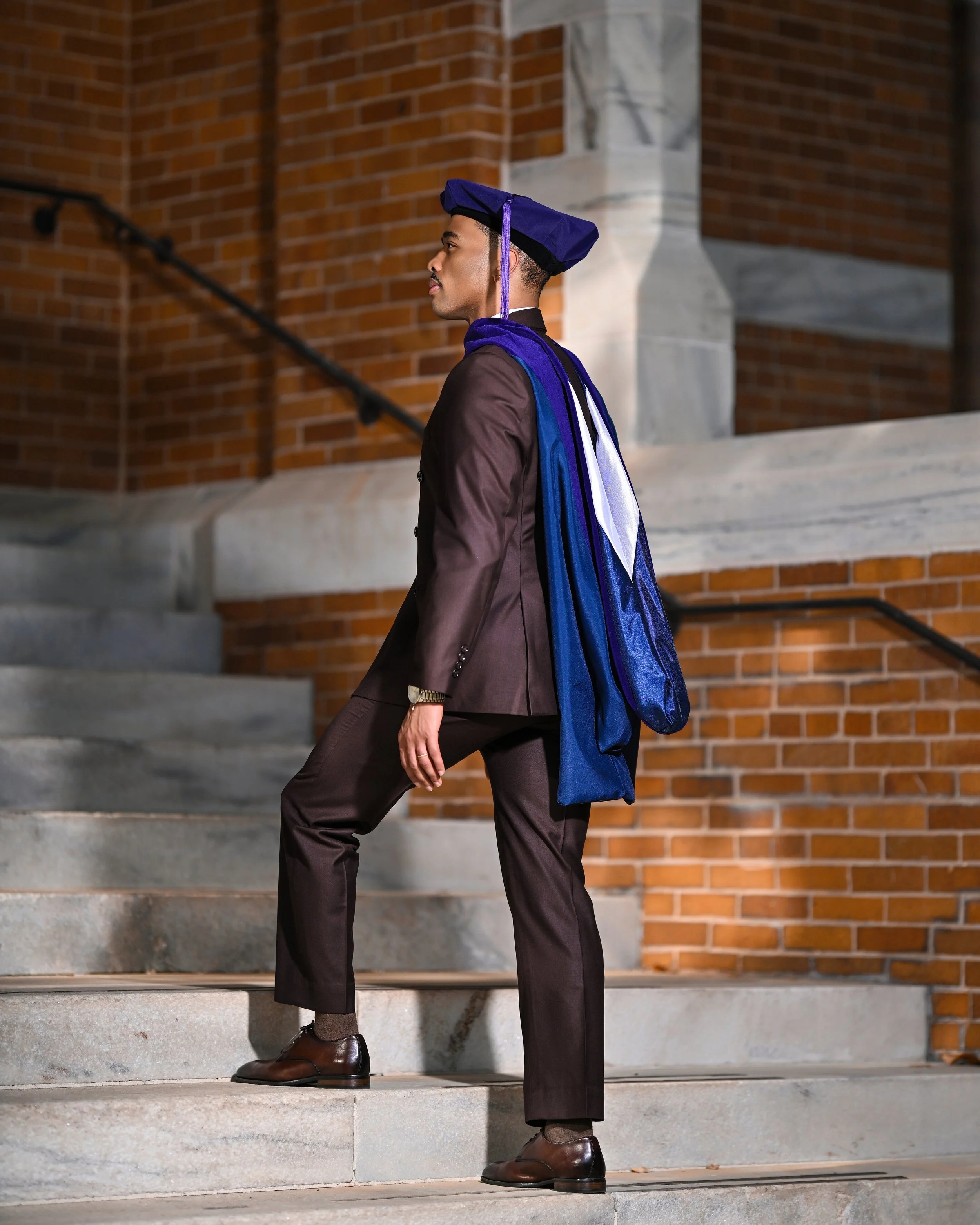 Man in formal attire with graduation cap and gown walking up stairs outside a brick building.