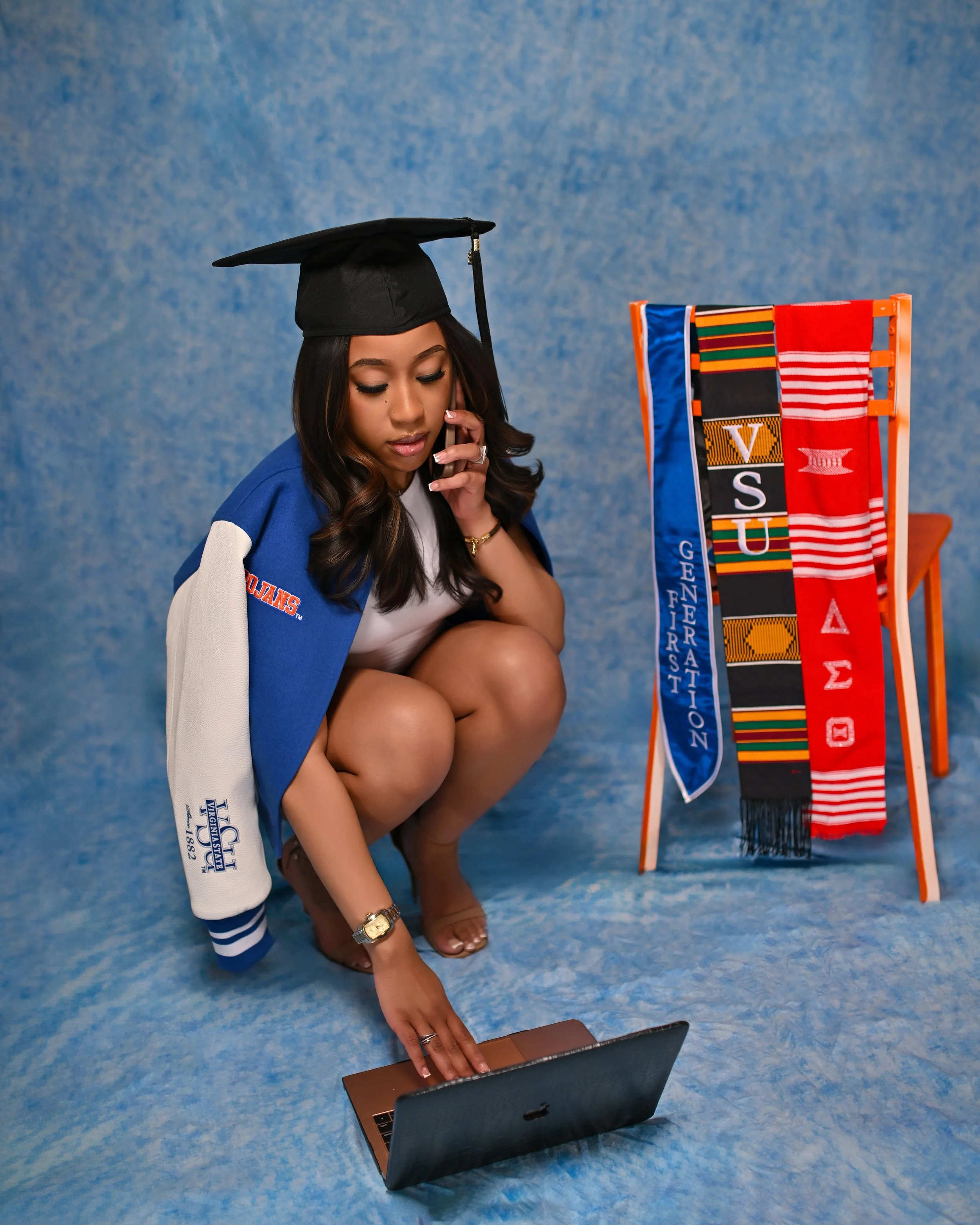 A young woman in a graduation cap and gown sitting on the floor, looking at a laptop, with a colorful display of graduation stoles or sashes beside her on a stand.