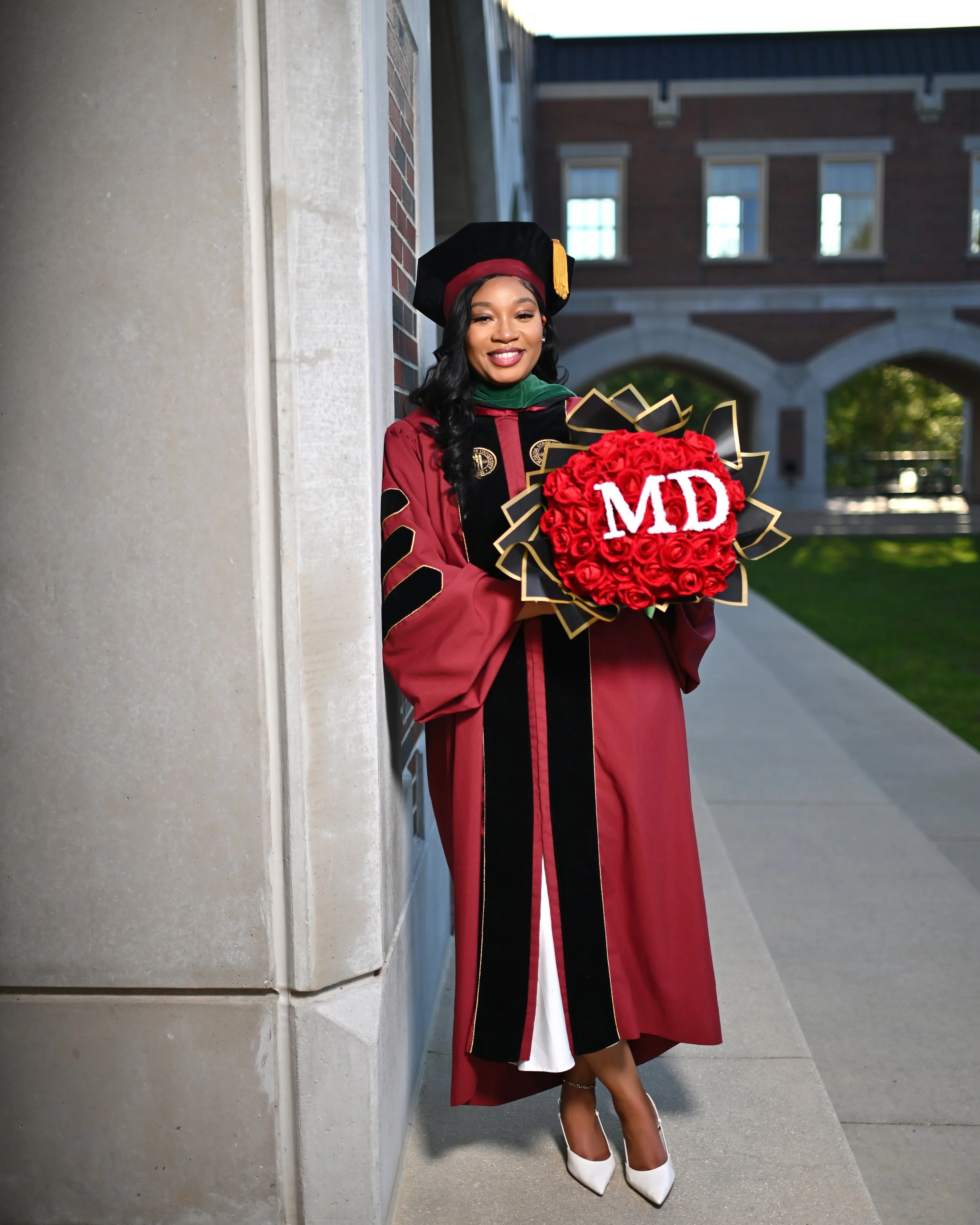 A young woman in a graduation cap and gown, holding a bouquet of red roses with 'MD' written on it, standing outdoors near a building with brick and stone walls, smiling.
