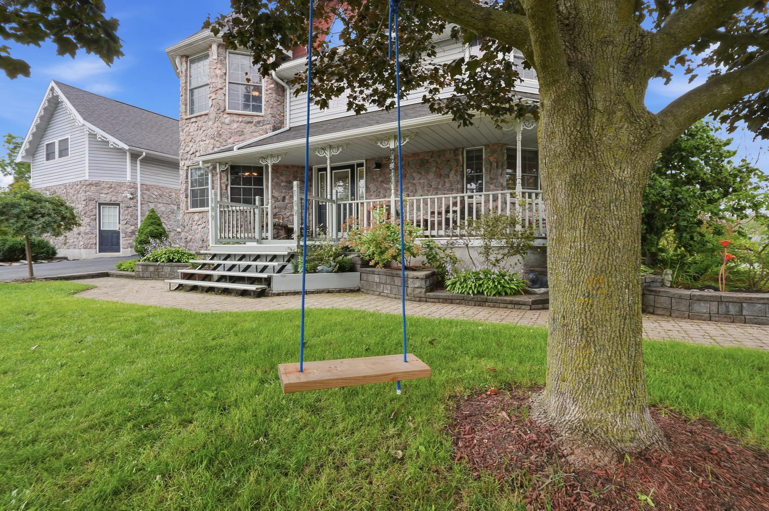 A backyard with a wooden swing hanging from a large tree, a brick house with a front porch, and well-maintained grass and plants.