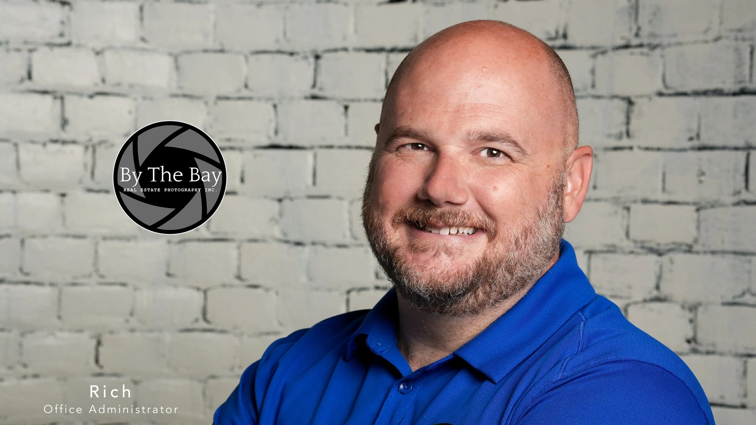 A headshot of a bald man with a beard, smiling in front of a white brick wall, wearing a blue collared shirt.