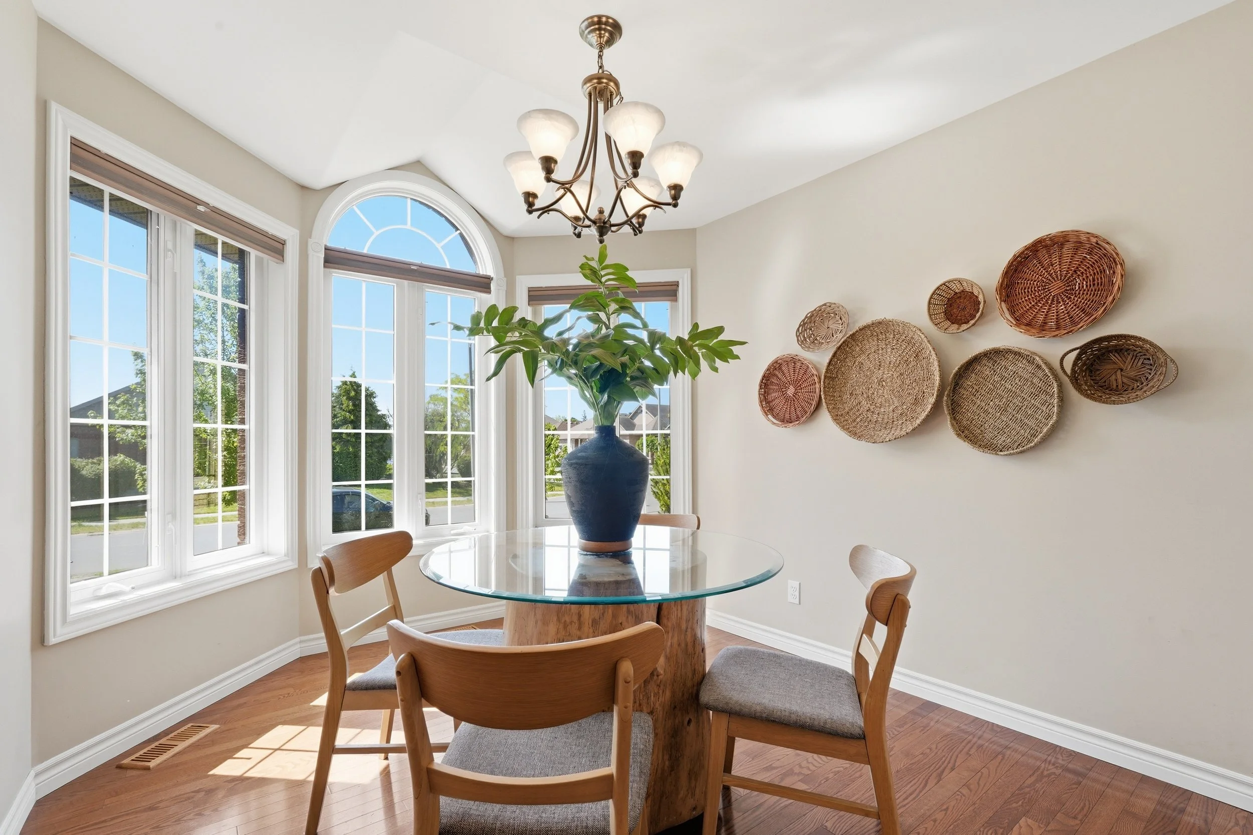 A dining area with a round glass table, surrounded by wooden chairs with fabric seats, a large vase with green foliage on the table, three large windows with white frames, a decorative wall hanging of woven baskets, and a chandelier with multiple gla
