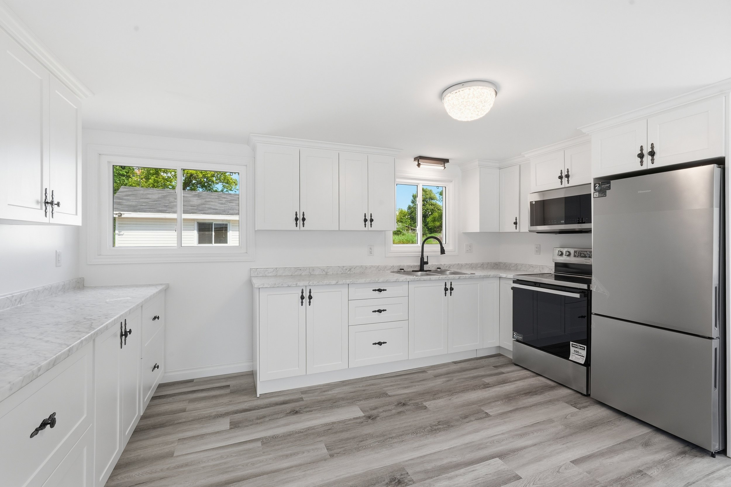 Bright white kitchen with white cabinets, marble countertops, stainless steel appliances, and large windows showing a view of a house and trees outside.