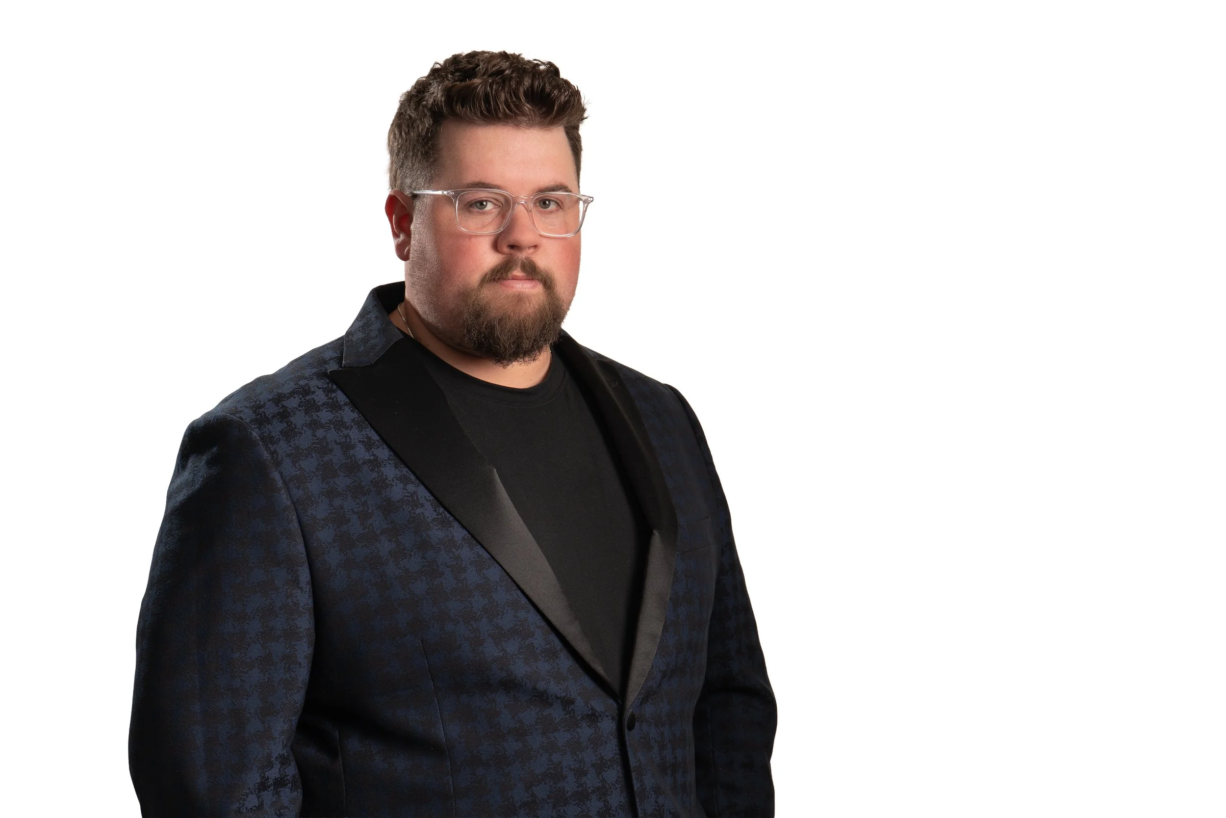 A man with glasses, a beard, and short brown hair wearing a black shirt and a navy blue blazer with a subtle pattern and satin lapel, standing against a plain white background.
