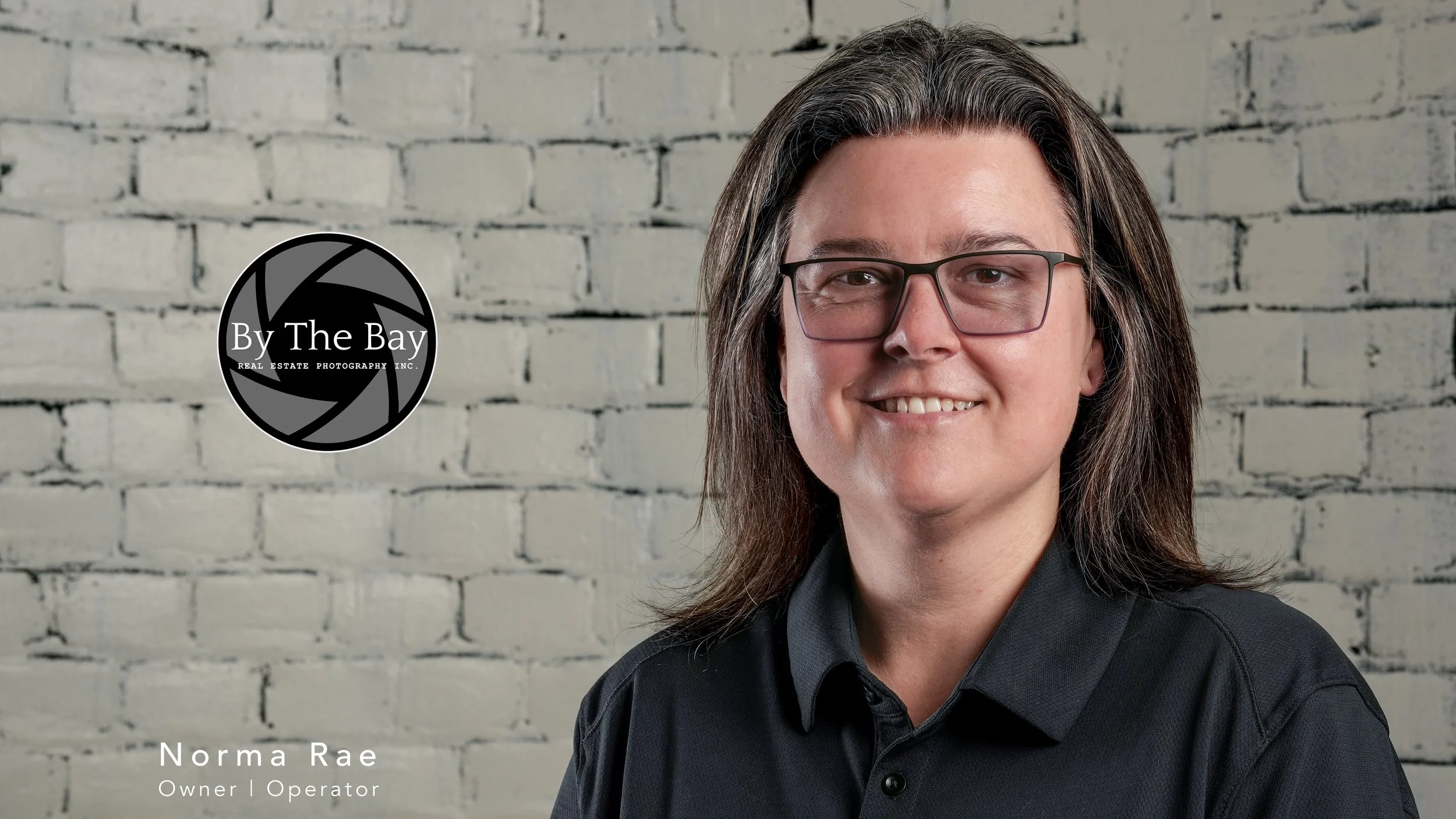 Headshot photo of Norma Rae, an owner and operator, smiling in front of a white brick wall. The image includes a logo of 'By The Bay Real Estate Photography Inc.' on the left side.