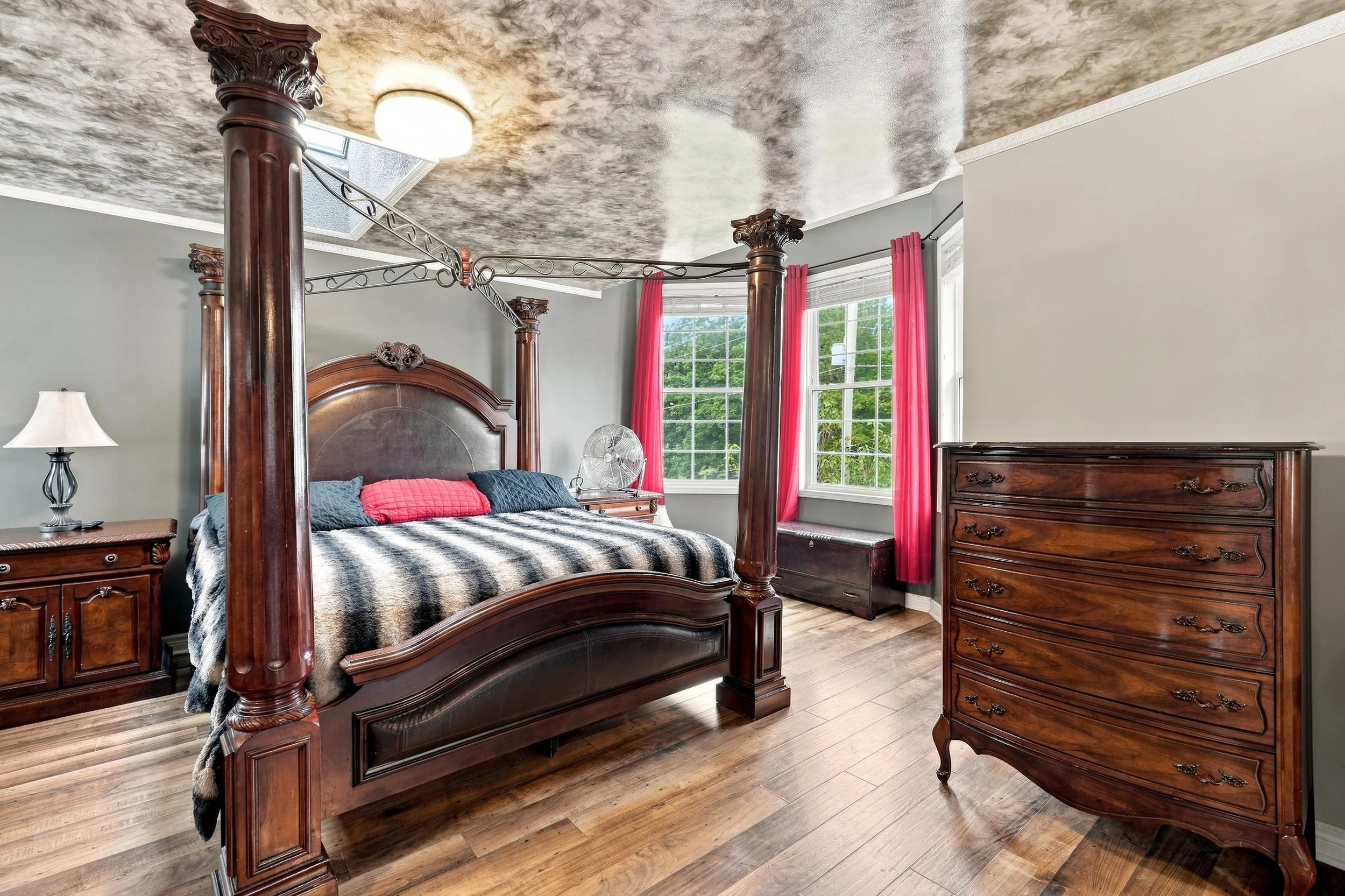A bedroom with a large wooden four-poster bed, a matching dresser, and a nightstand with a lamp. The room has gray walls, a window with red curtains, and a textured ceiling.