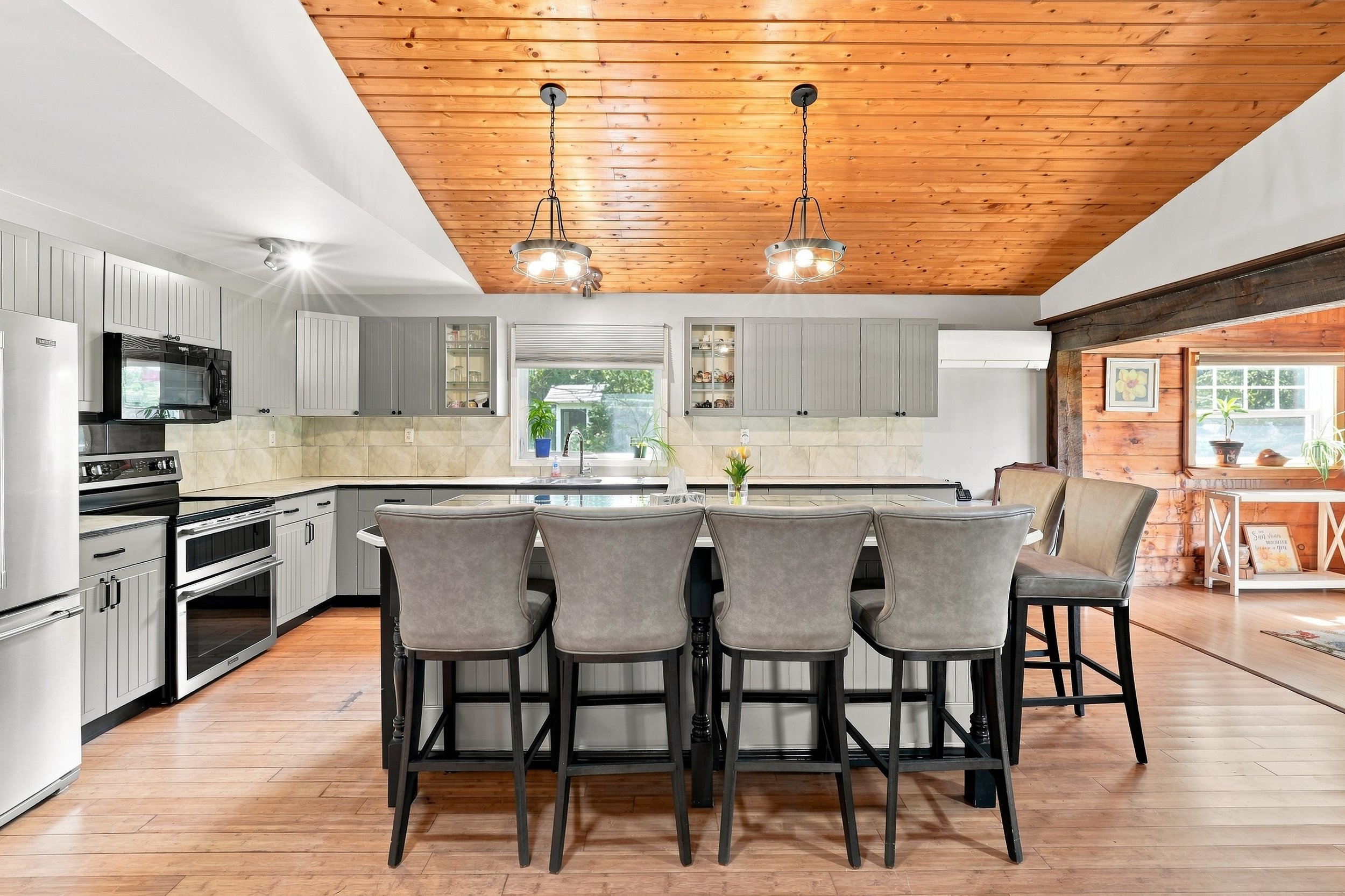 View of a modern kitchen and dining area with a wooden vaulted ceiling, pendant lights, white cabinets, a window with plants, and a kitchen island with chairs.