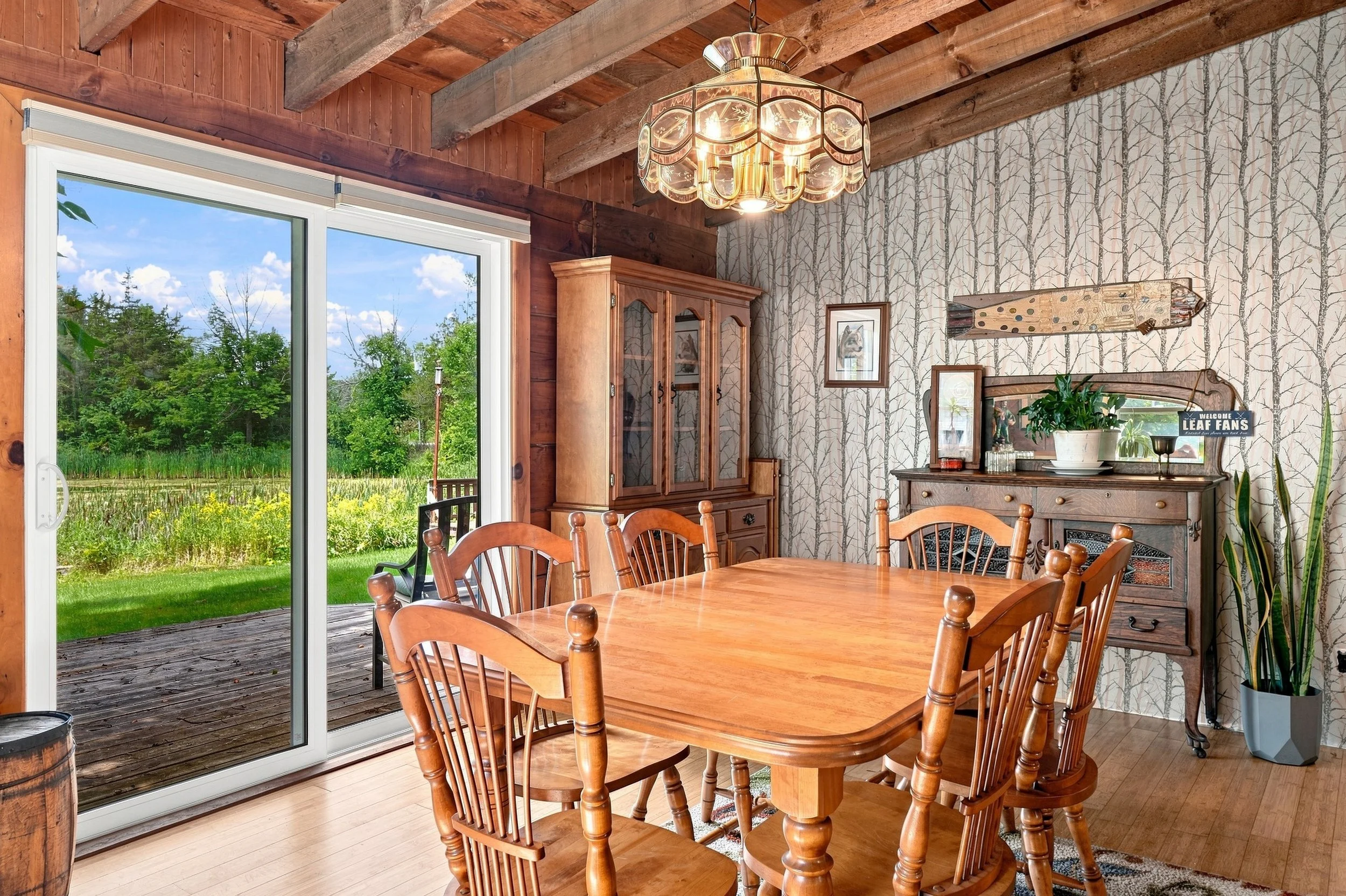 A dining room with a wooden table and chairs, a china cabinet, and a fireplace, with a sliding glass door that opens to a deck overlooking a green yard and trees.