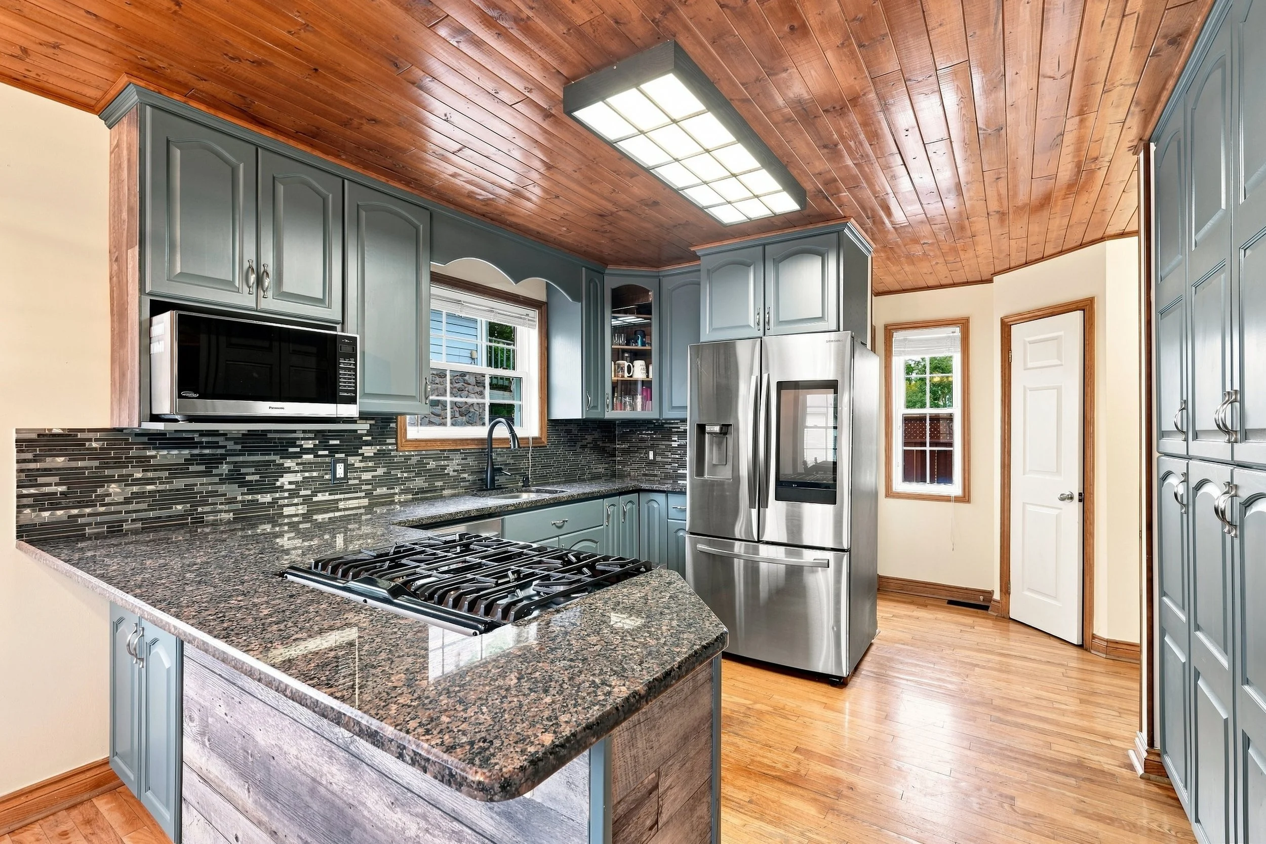 Kitchen with blue cabinets, granite countertop island, stainless steel refrigerator, microwave, gas stove, wood ceiling and hardwood floors, windows with natural light.