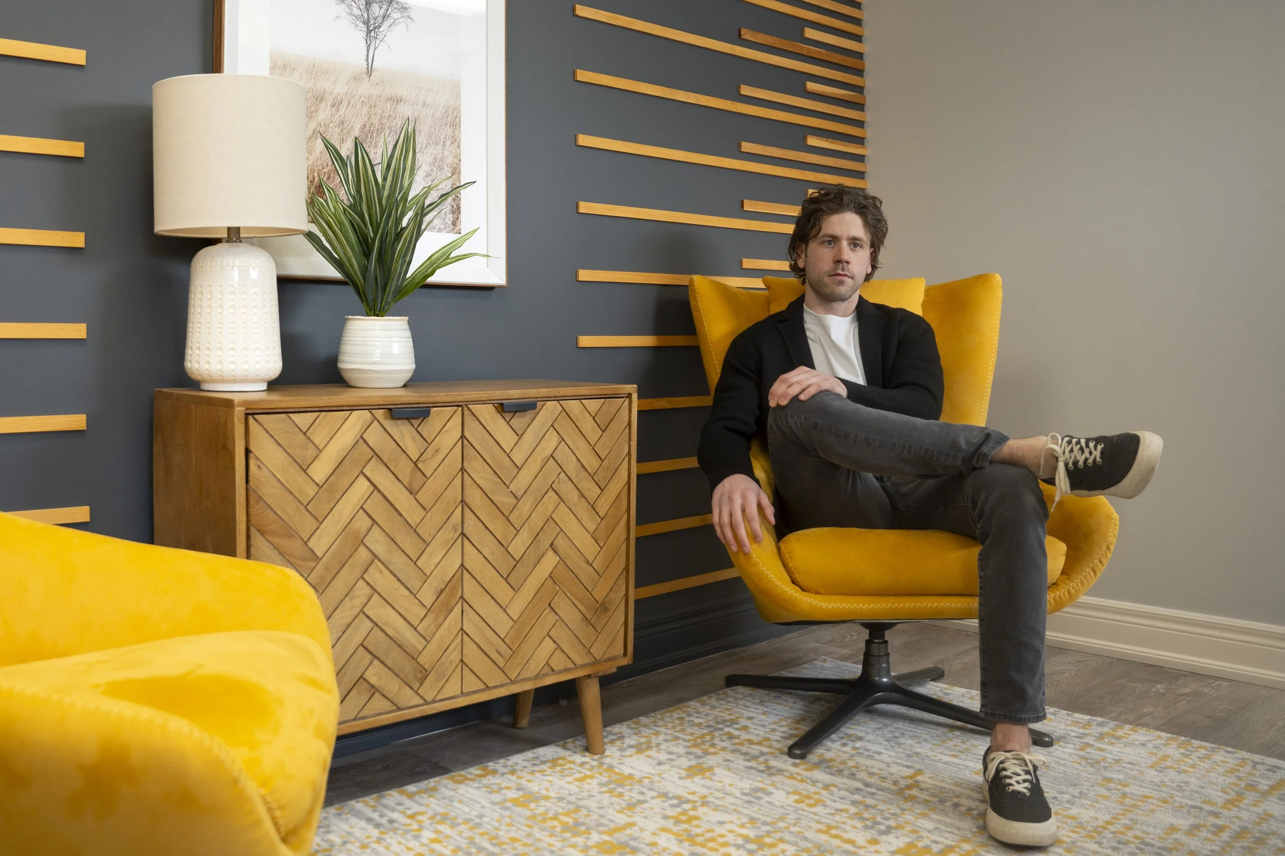 A man sitting in a yellow armchair in a modern living room with gray and wooden accents, a sideboard with a white lamp and potted plant, and abstract wall art.