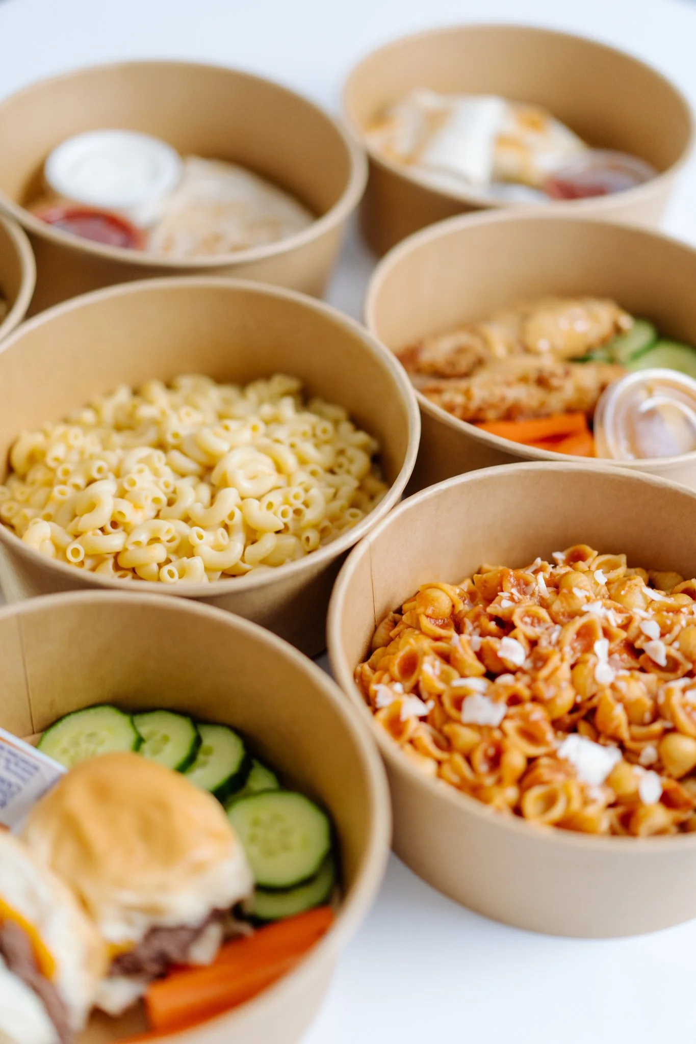 Several brown paper bowls containing different types of pasta, vegetables, and sauces, with one bowl showing sliced cucumbers and a sandwich bun.