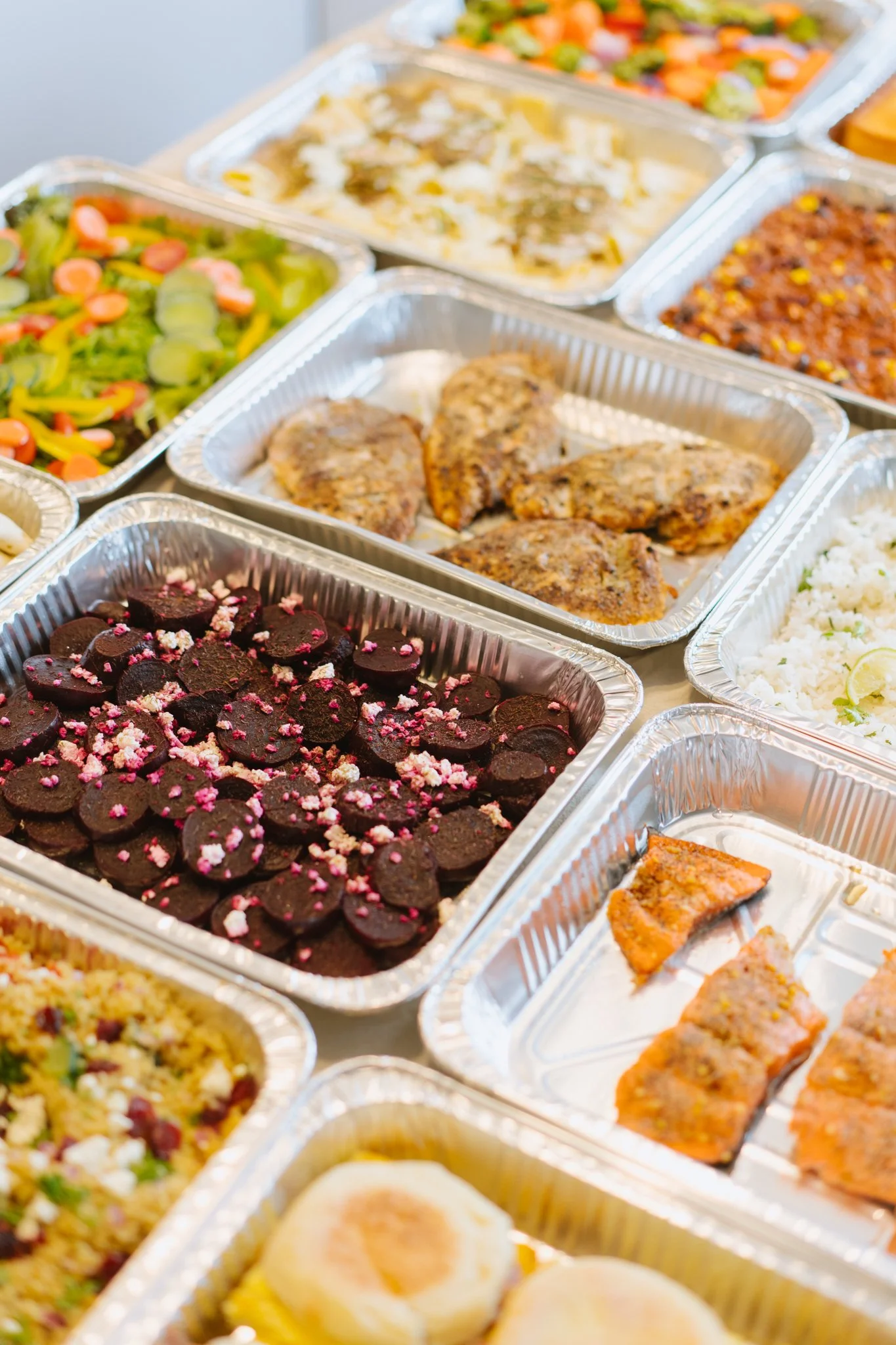 Assorted takeout food containers filled with various dishes, including fried chicken, salad, rice, pasta, sliced meatloaf, and chocolate cookies with pink sprinkles.