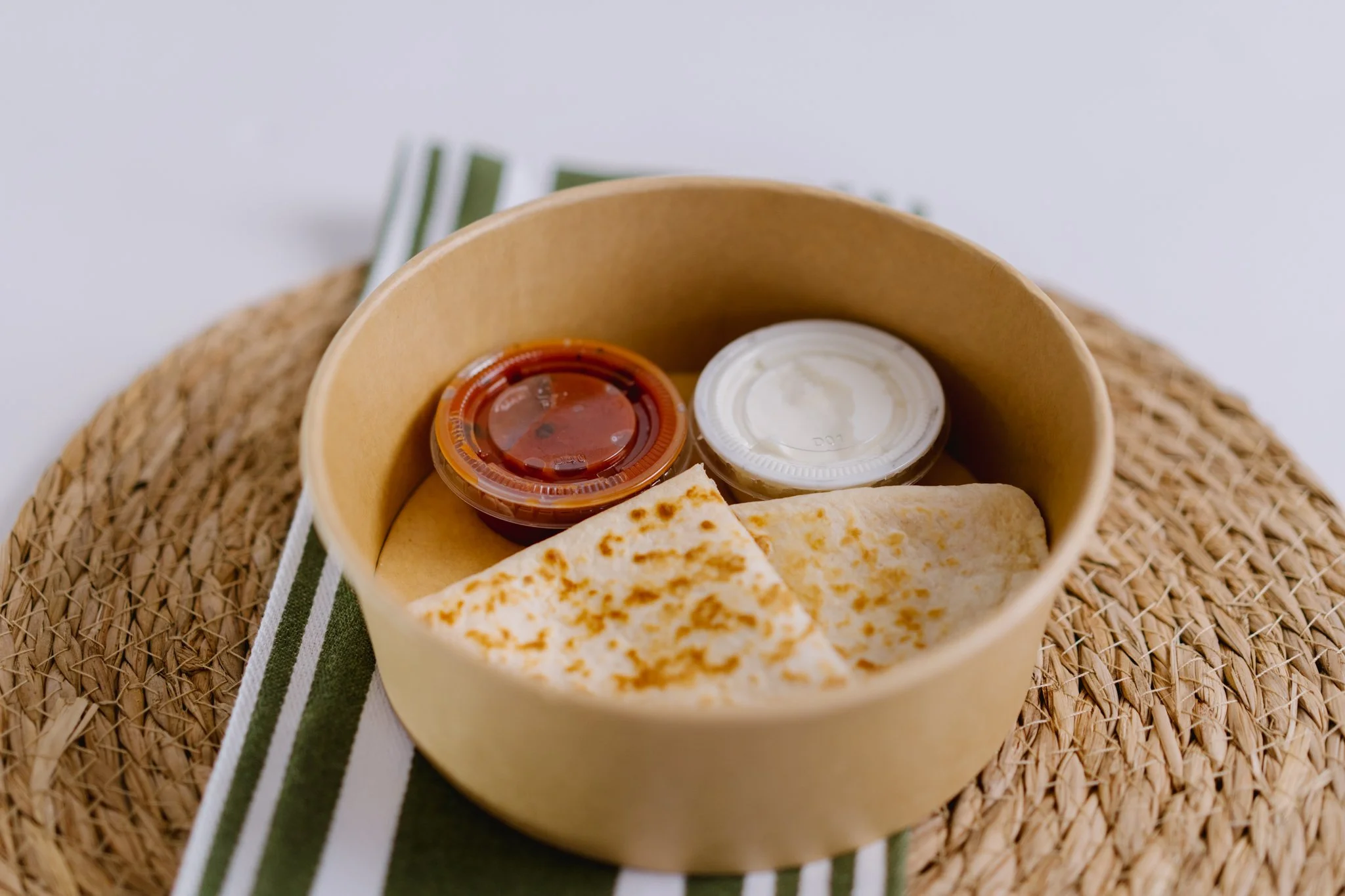 A round cardboard container holds two cups of sauce and a folded flatbread, placed on a woven placemat with a striped cloth underneath.
