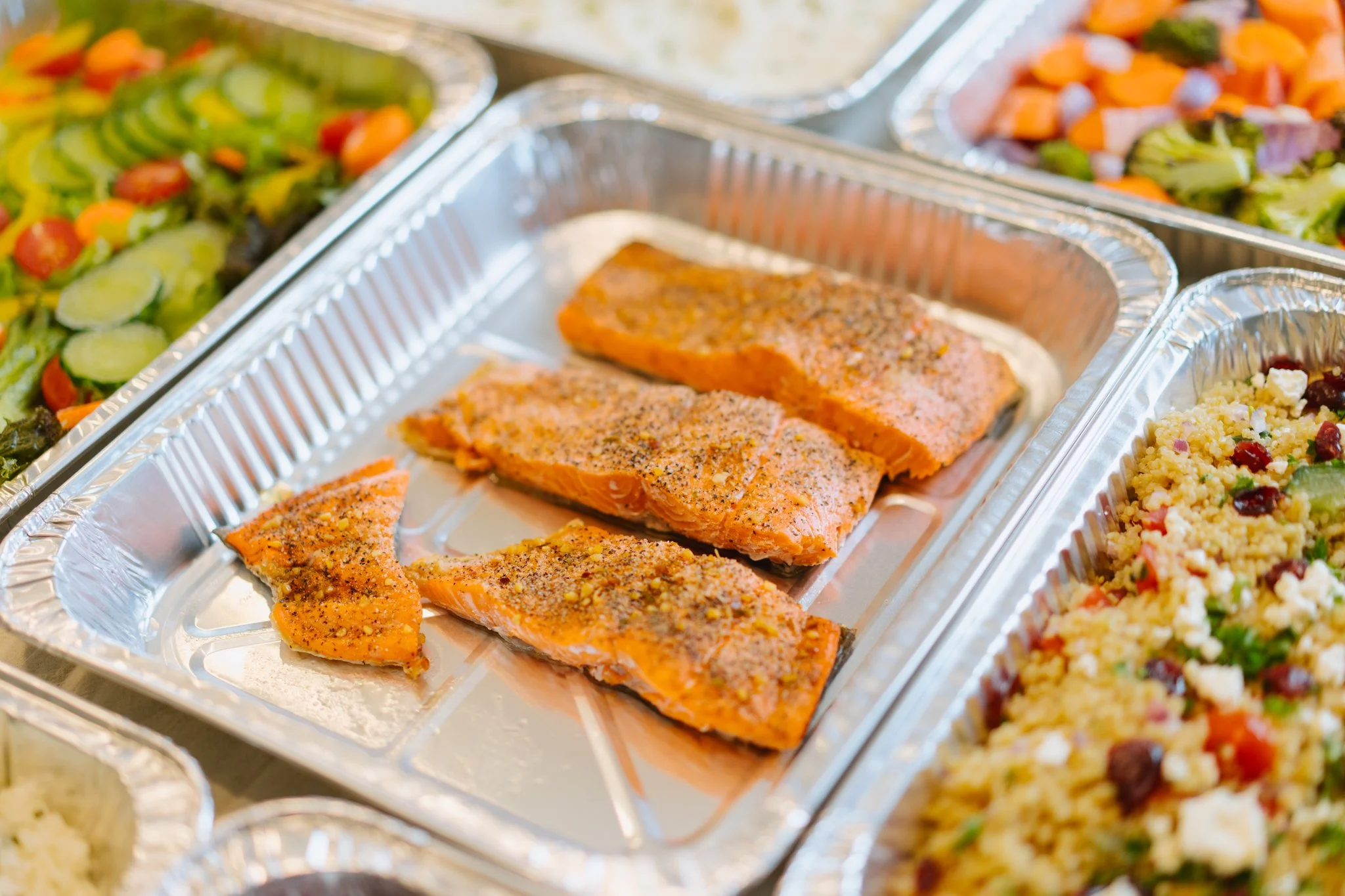Baked salmon fillets seasoned with black pepper in a foil tray, surrounded by salads with vegetables.