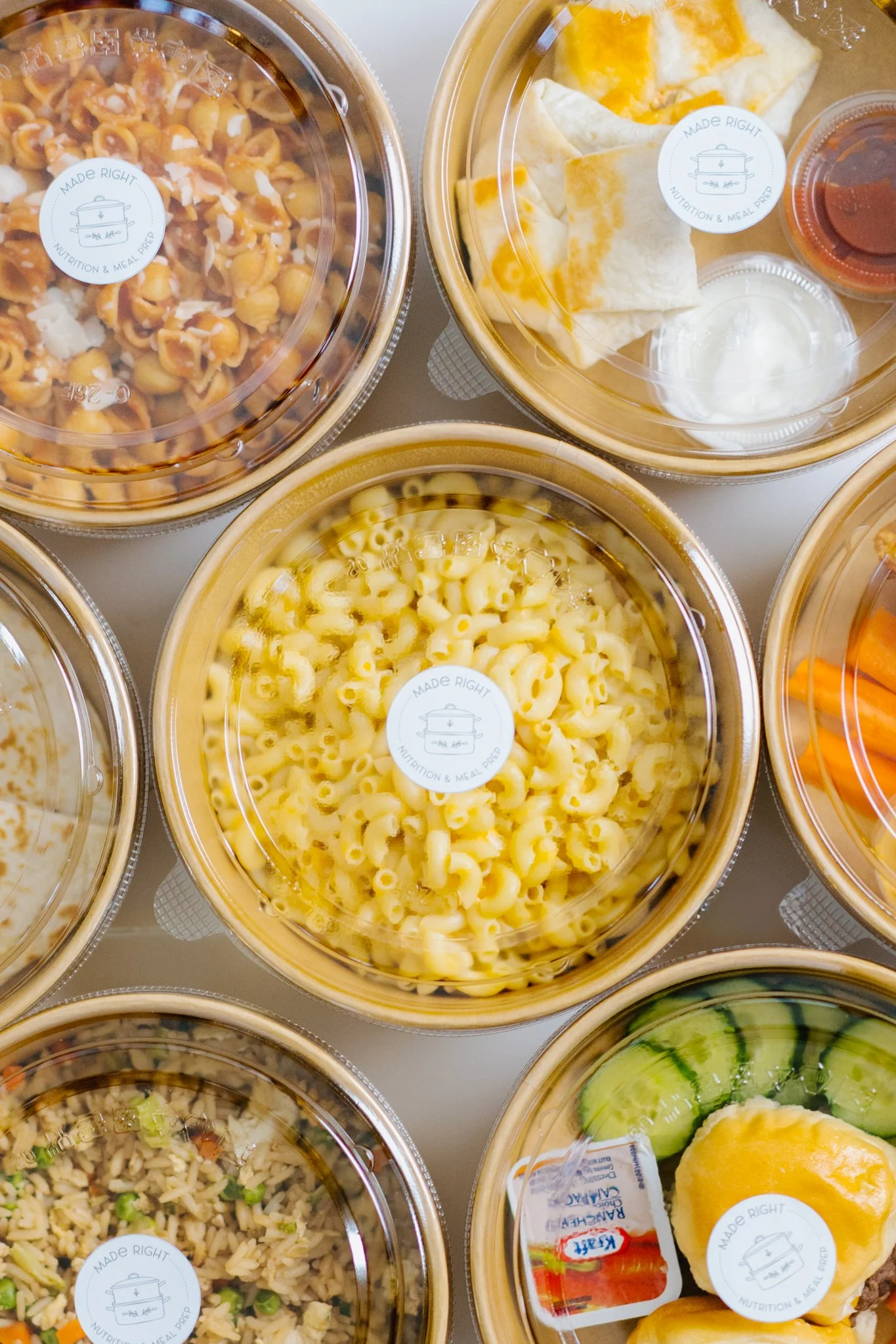 Assorted pre-packaged meals in plastic containers, including macaroni and cheese, fried rice, pasta salad, and grilled vegetables, with some containers containing dipping sauces or condiments.