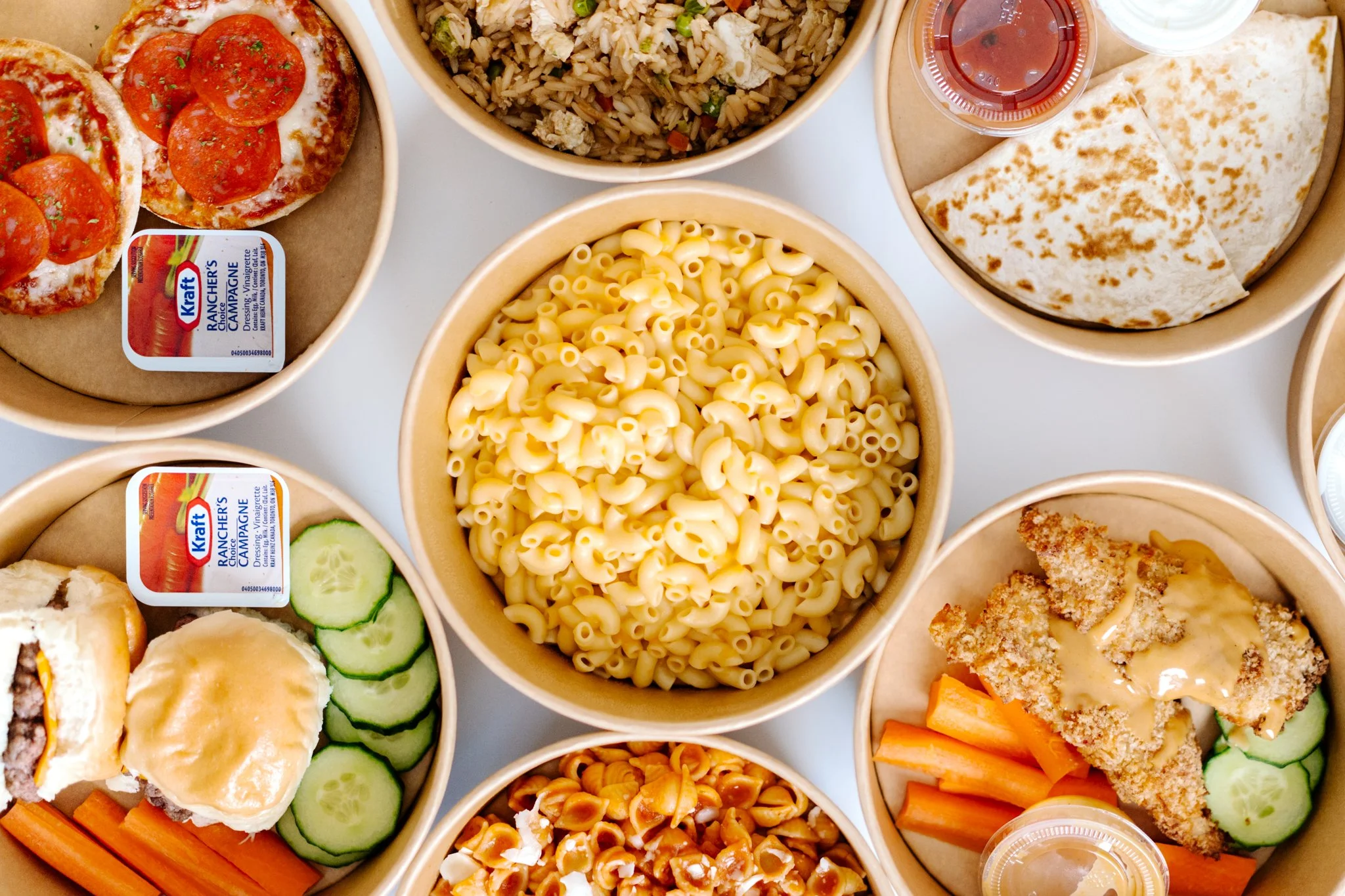 Downward view of various bowls of food, including pasta, pizza, fried chicken, carrots, cucumber slices, and a sandwich with a side of ranch dressing.
