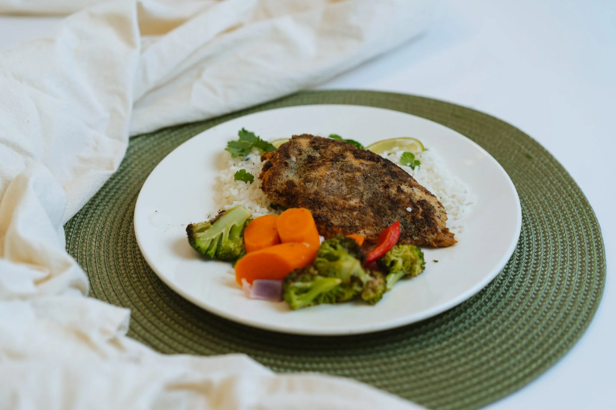 A white plate with rice, a fried fish fillet, and mixed vegetables including broccoli, carrots, red pepper, and onion, on a green woven placemat with a cream-colored cloth nearby.