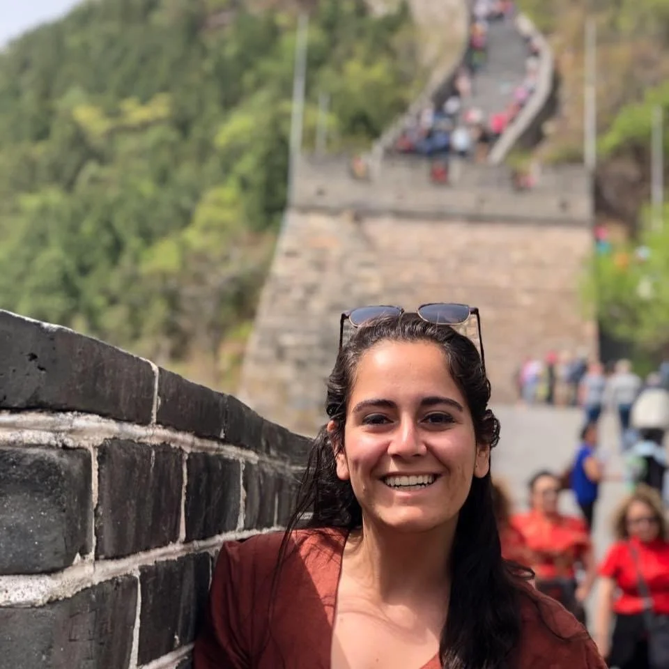 Smiling young woman with dark hair, wearing sunglasses on her head, standing next to a stone wall at a popular tourist site. The Great Wall of China is visible in the background with visitors walking along it.