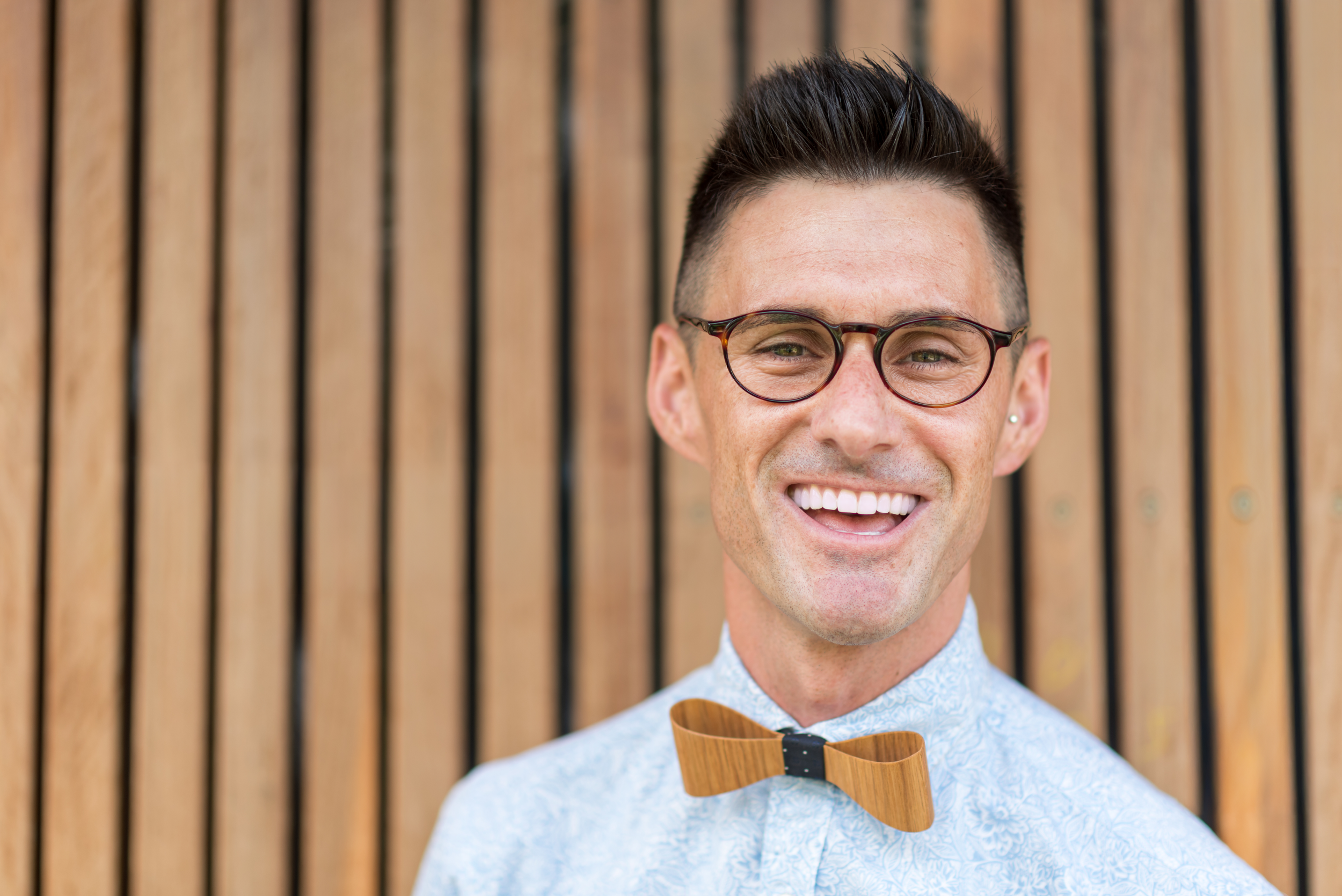 A smiling man with glasses and a wooden bow tie standing in front of a wooden slat wall.