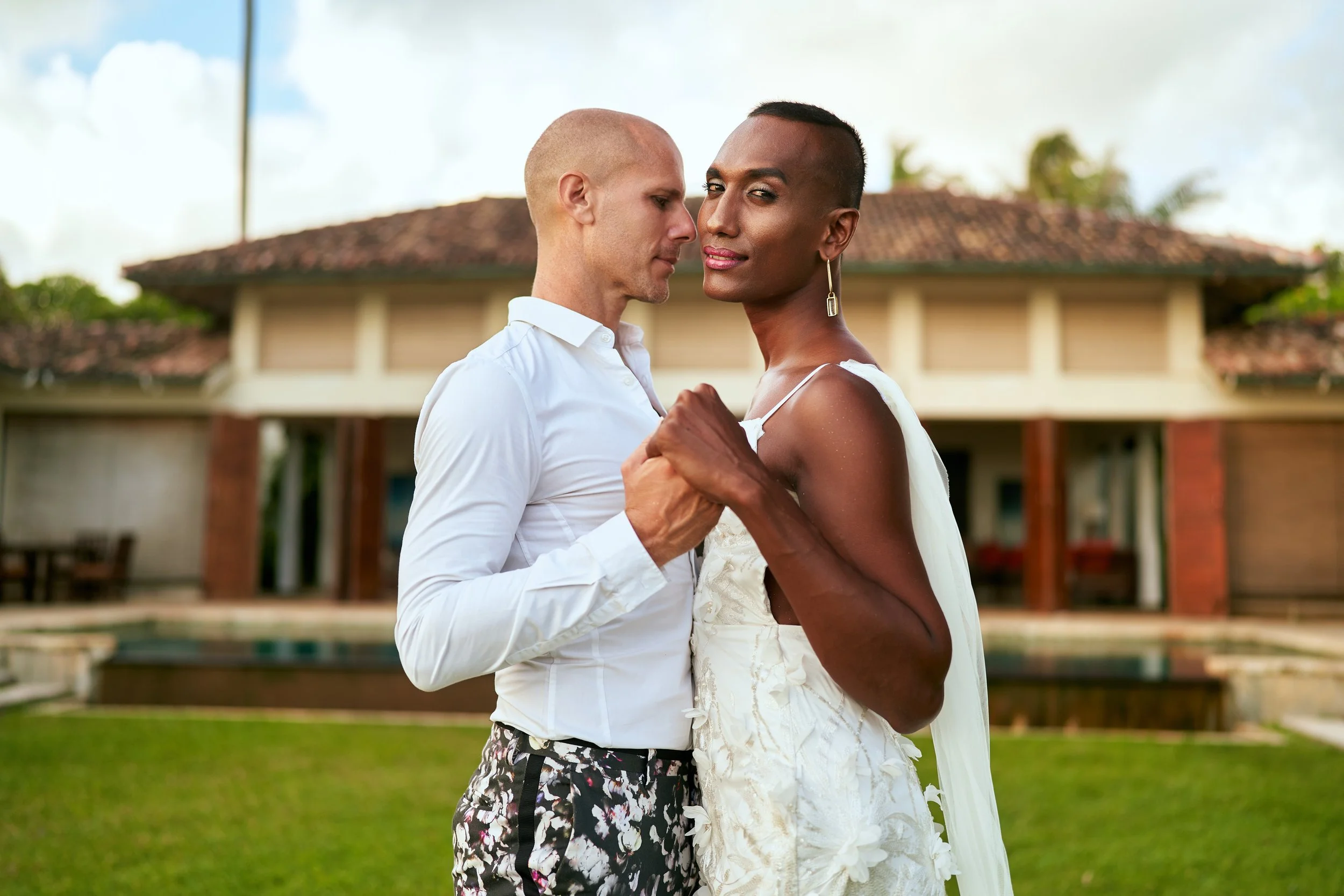 A couple dancing outdoors in front of a house, with greenery and a fountain in the background.