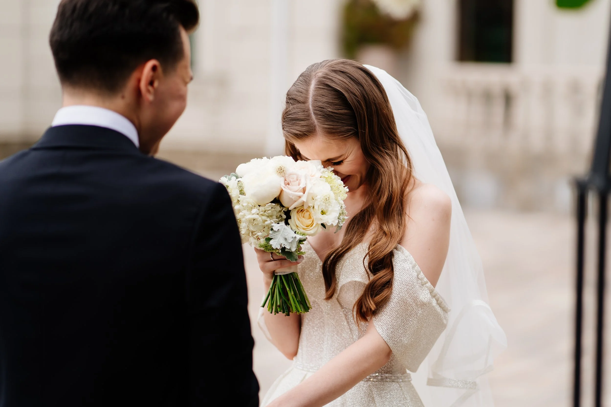 A bride with long, wavy brown hair, holding a bouquet of white and pale pink flowers, tilting her head and smiling while looking down, during a wedding ceremony with a groom in a black suit standing nearby.