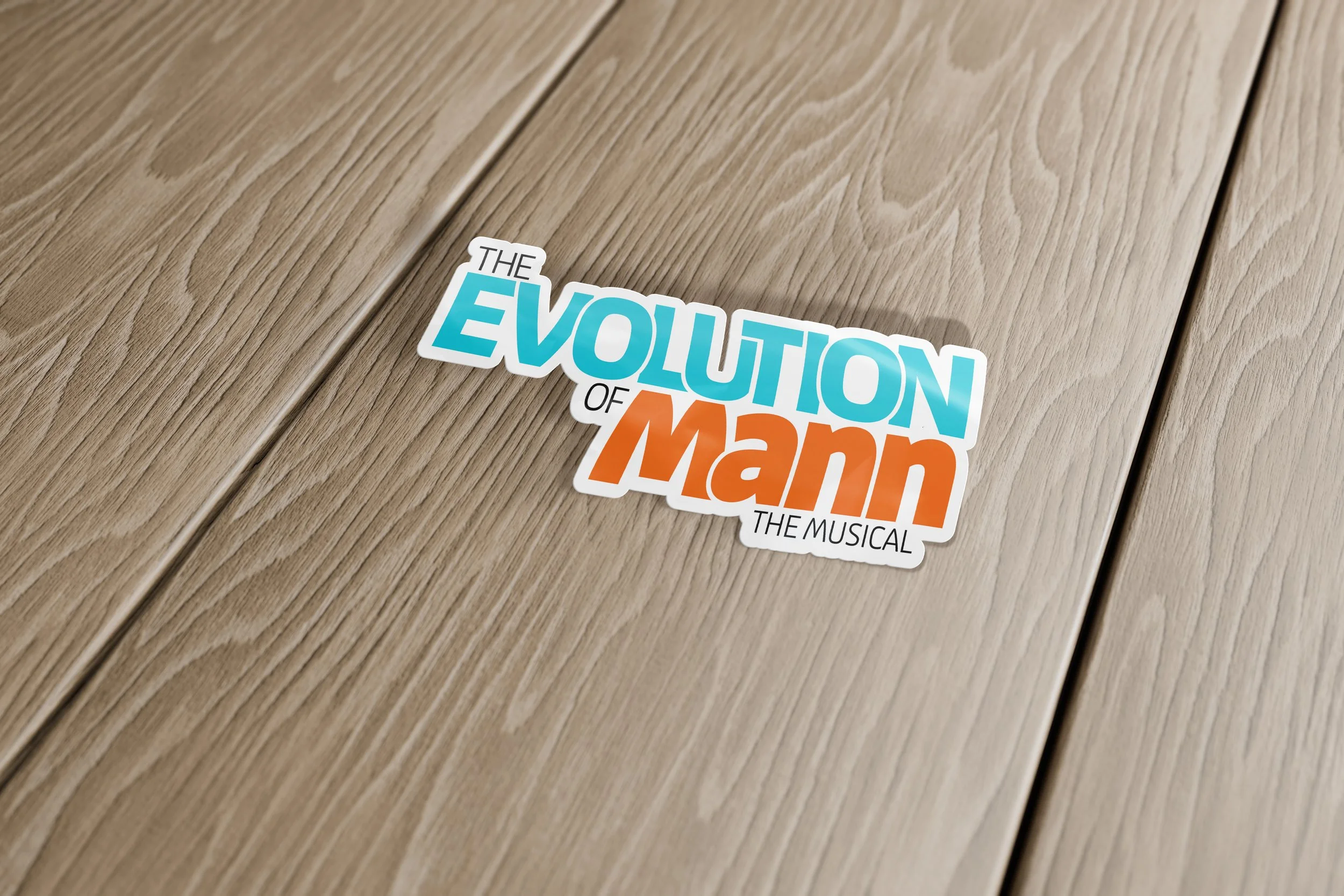 Sticker of the title 'The Evolution of Man The Musical' on a light wooden surface.