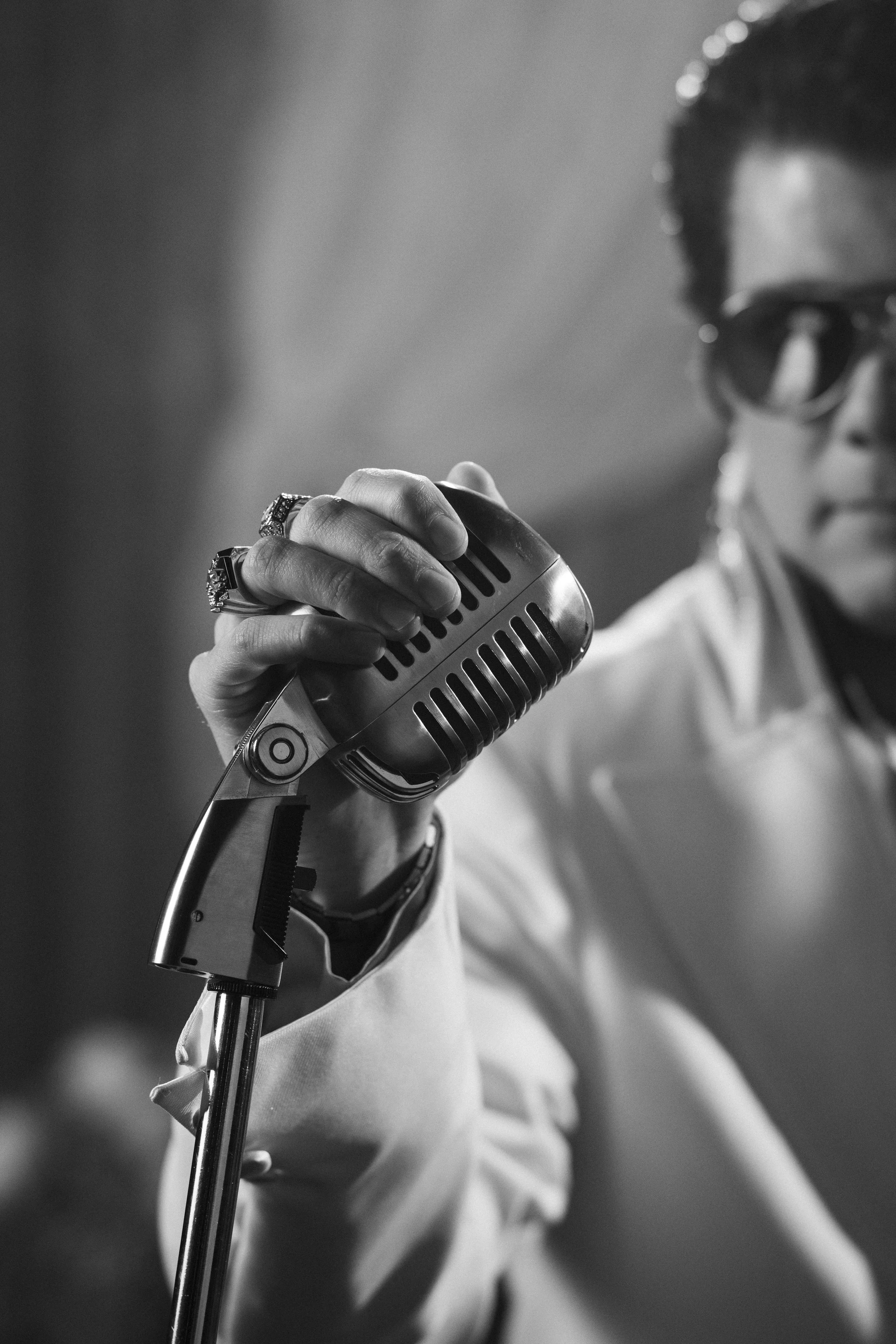 Black and white photo of a person holding a vintage microphone close to their mouth, wearing sunglasses and a light-colored shirt.