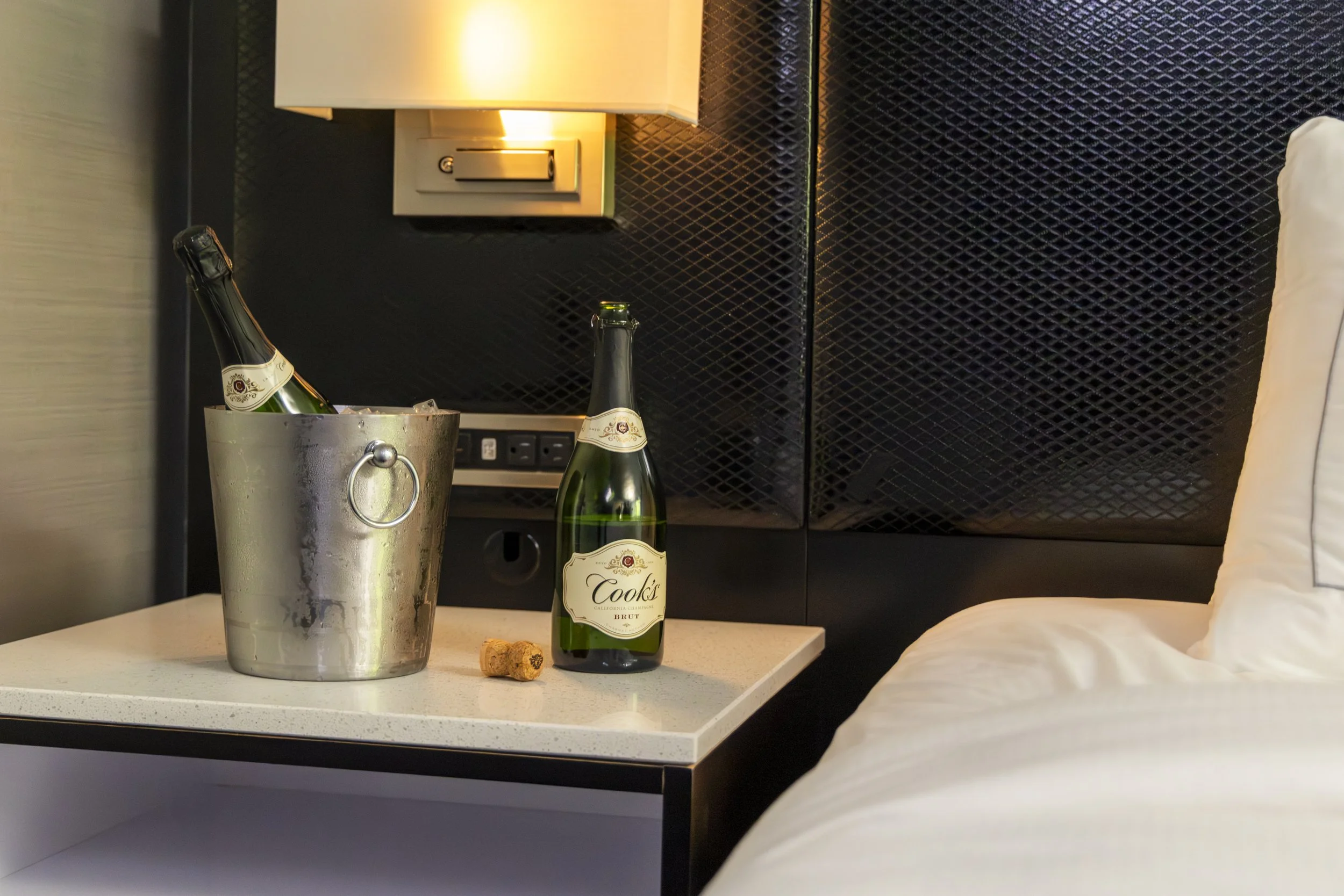 A bedside table with a silver champagne bucket containing two bottles of champagne, a bottle of Cook's champagne, and a wine cork, next to a bed with white bedding, against a black textured wall with a wall-mounted lamp.