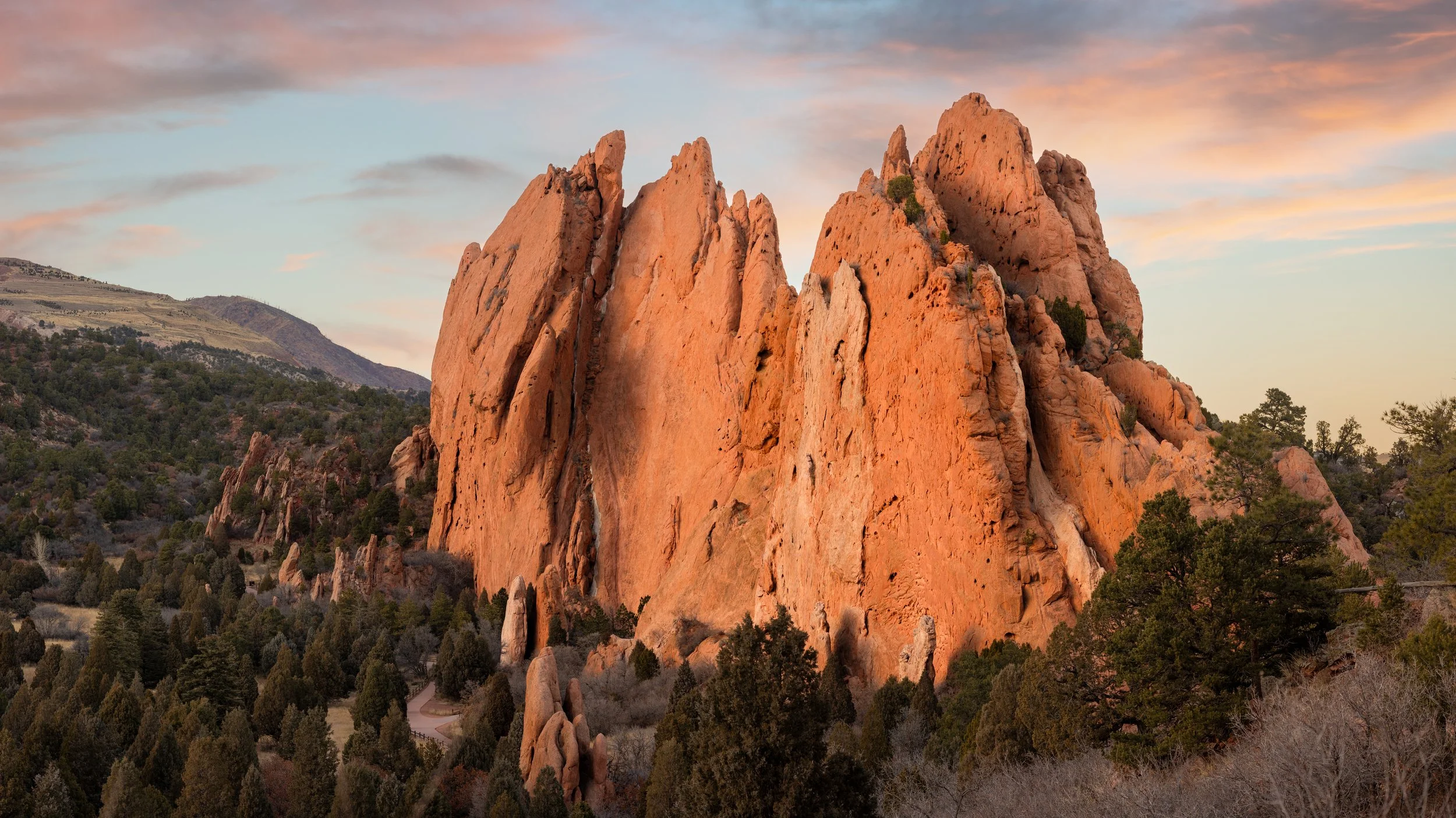 Pinkish-orange rock formations with green trees and a winding dirt path at sunrise or sunset.