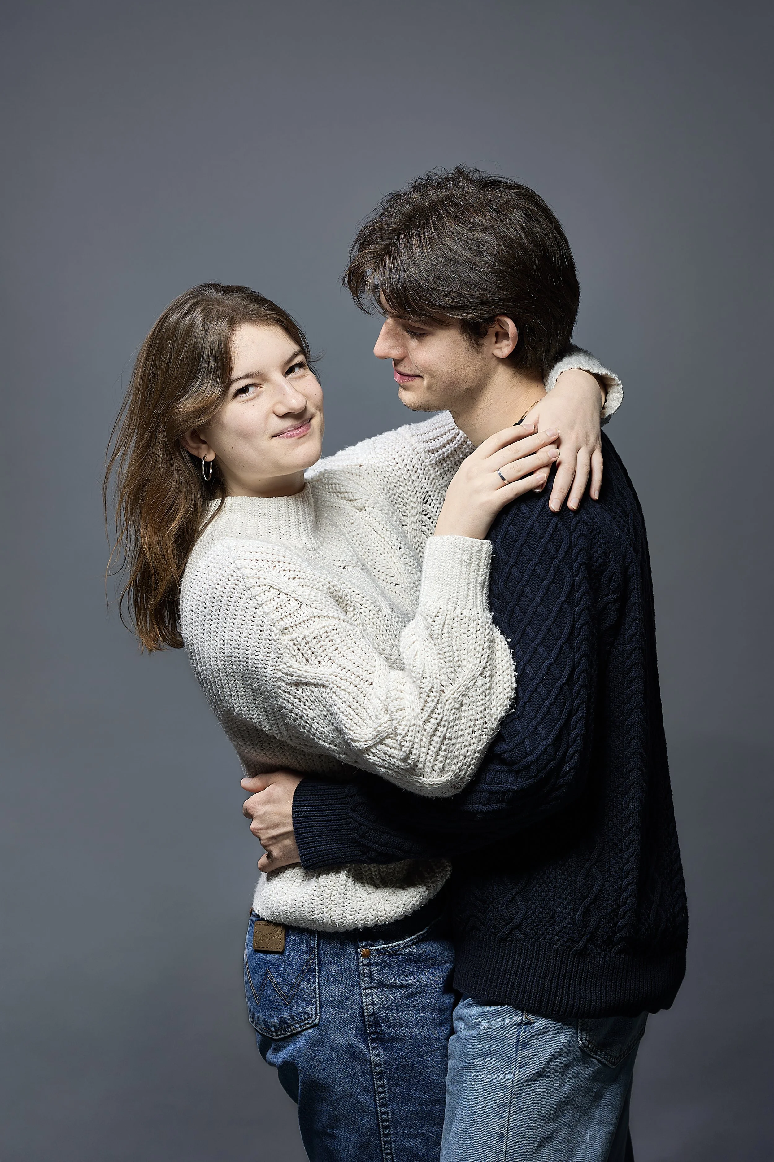 Young couple embracing, the woman with long hair wearing a white sweater, and the man with dark hair wearing a dark sweater, standing against a gray background.