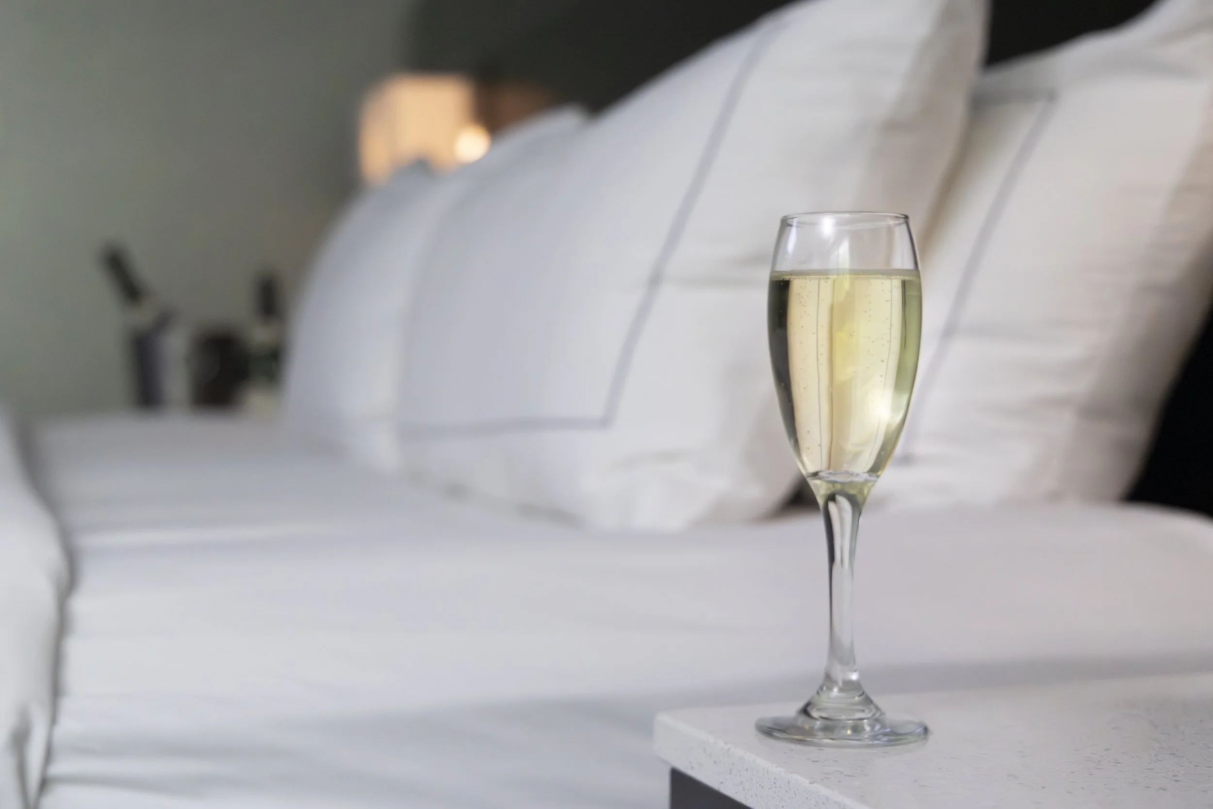 A glass of champagne on a nightstand next to a bed with white bedding in a cozy bedroom.